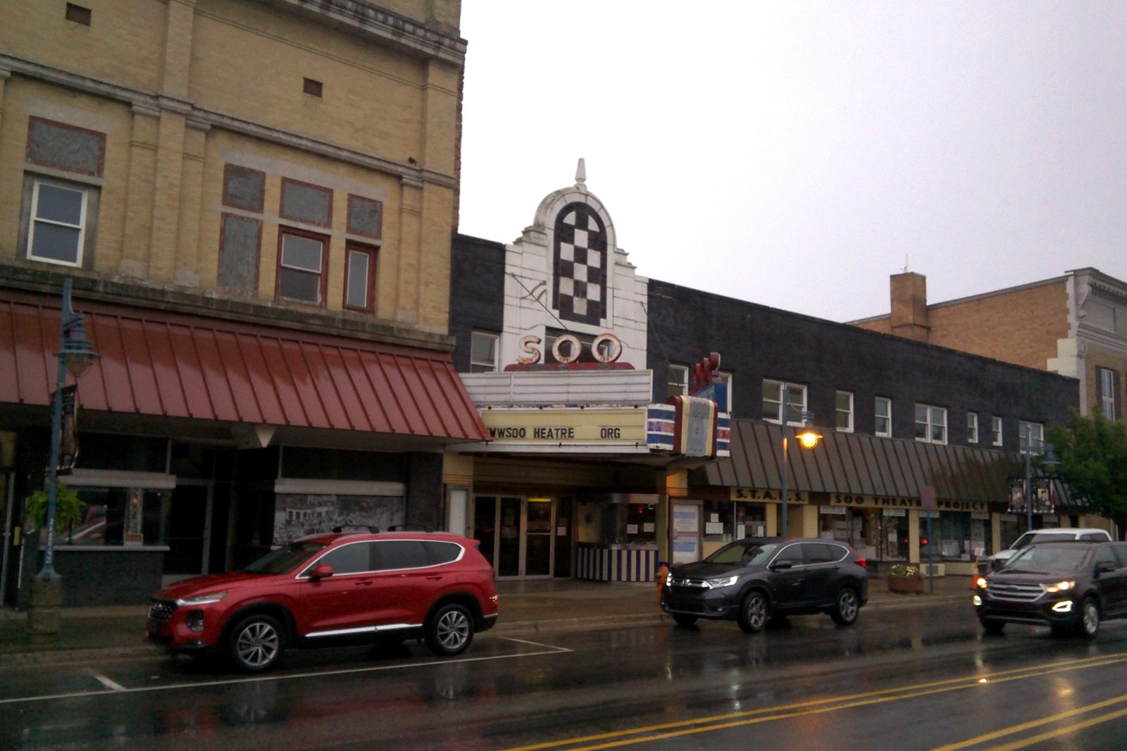 A downtown theater with a tall, ornate white-and-black sign tower over its entrance, between brick storefronts on a wet street with cars passing.