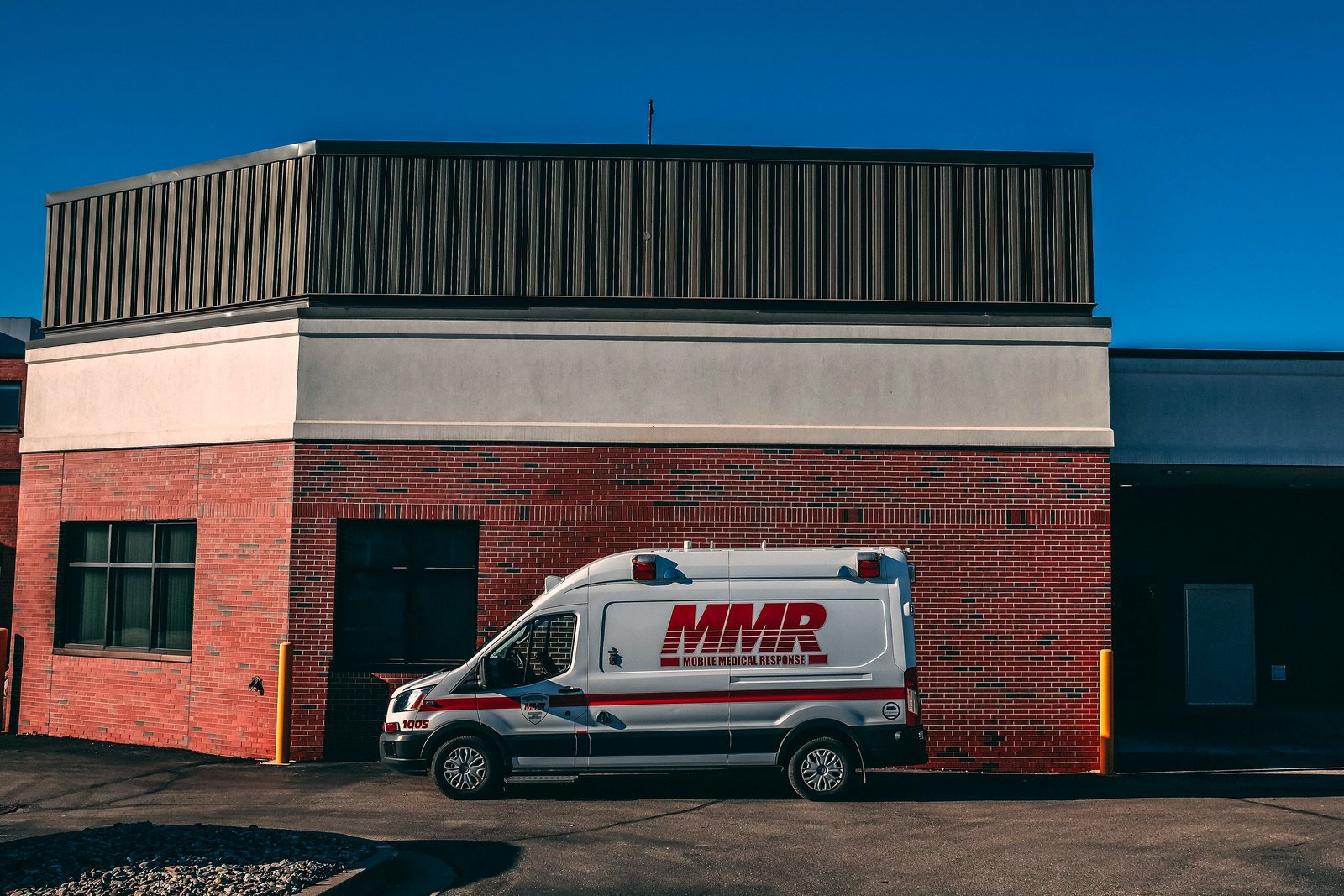 A white ambulance with red stripes and MMR lettering parked beside a red brick building with a tan upper wall, under a deep blue sky.