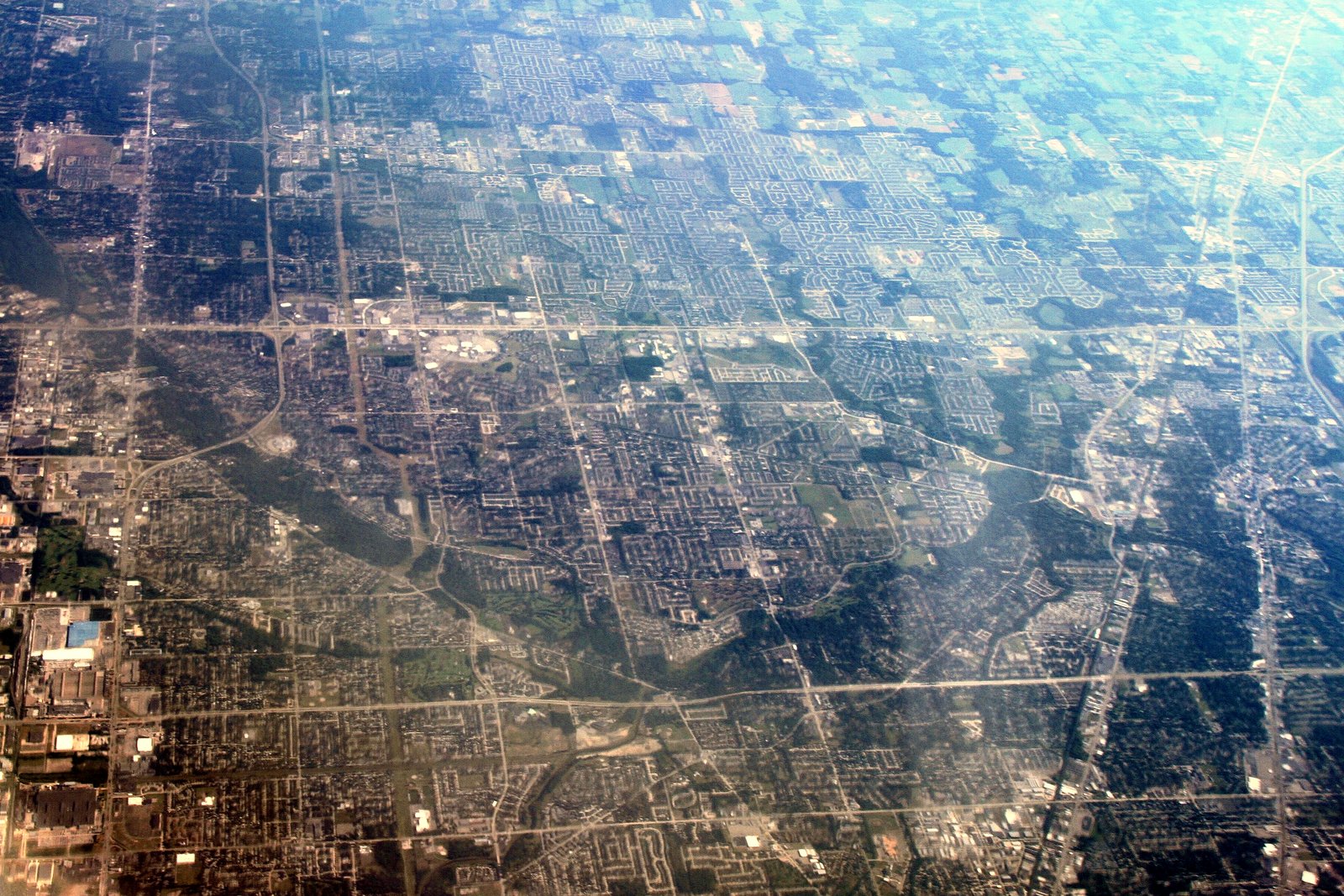 A hazy aerial view of a flat suburban grid of neighborhoods and roads, with a dark wooded river corridor curving through the middle.