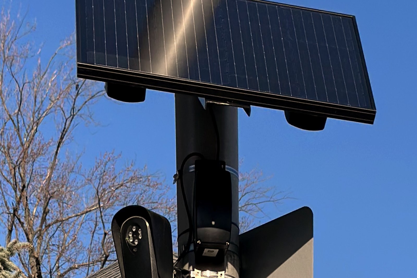 A small black camera mounted on a metal pole under a square solar panel, against a blue sky with bare trees.