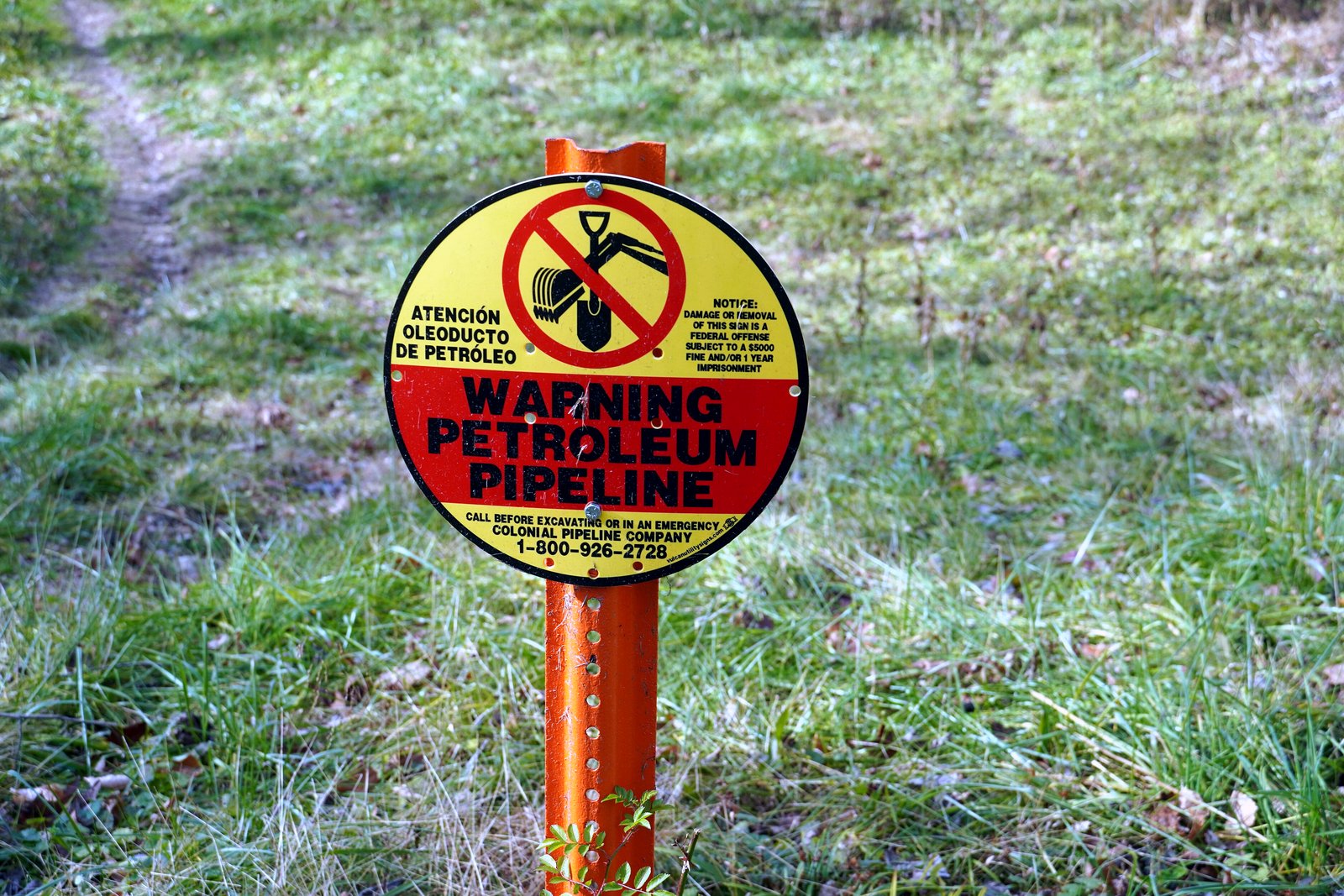 A round yellow and red sign reading Warning Petroleum Pipeline on an orange post in a grassy field.