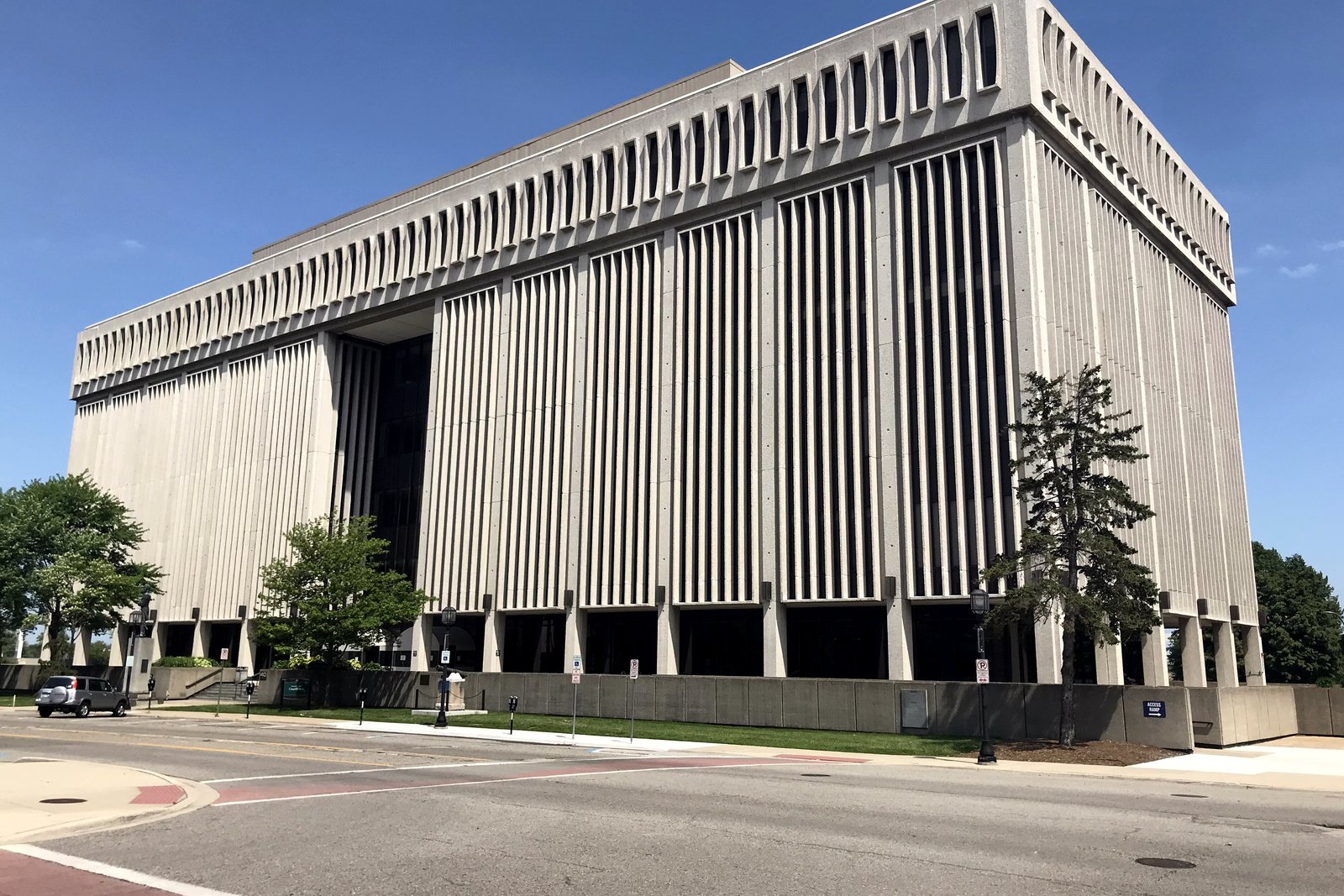 A tall, pale concrete office tower with vertical ribs and a recessed ground floor stands beside a street corner under a blue sky.