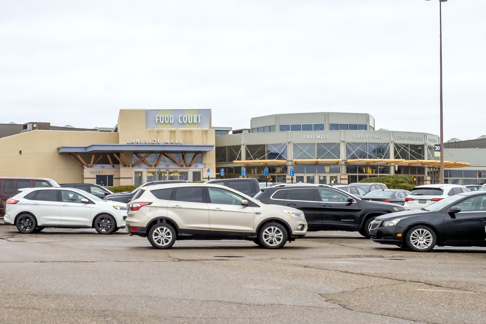 A large shopping mall with a tan and red facade behind a wide parking lot filled with cars on an overcast day.