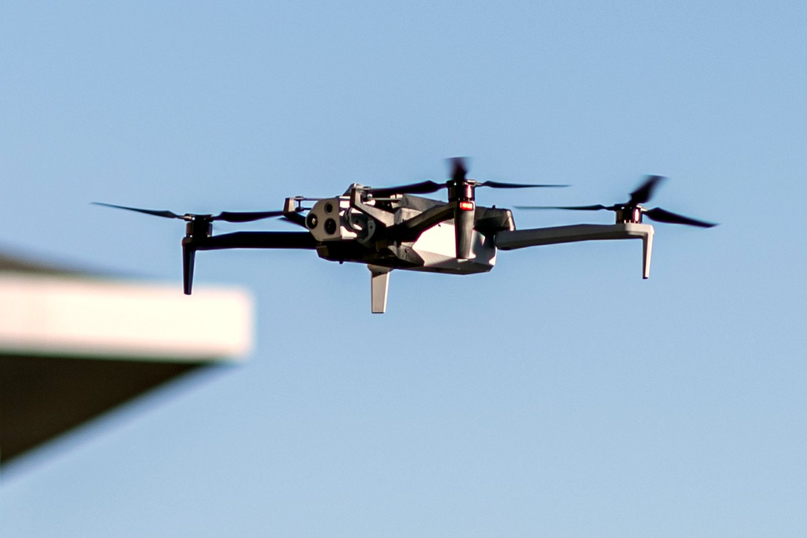 A dark gray quadcopter drone with four rotors flying against a clear blue sky.
