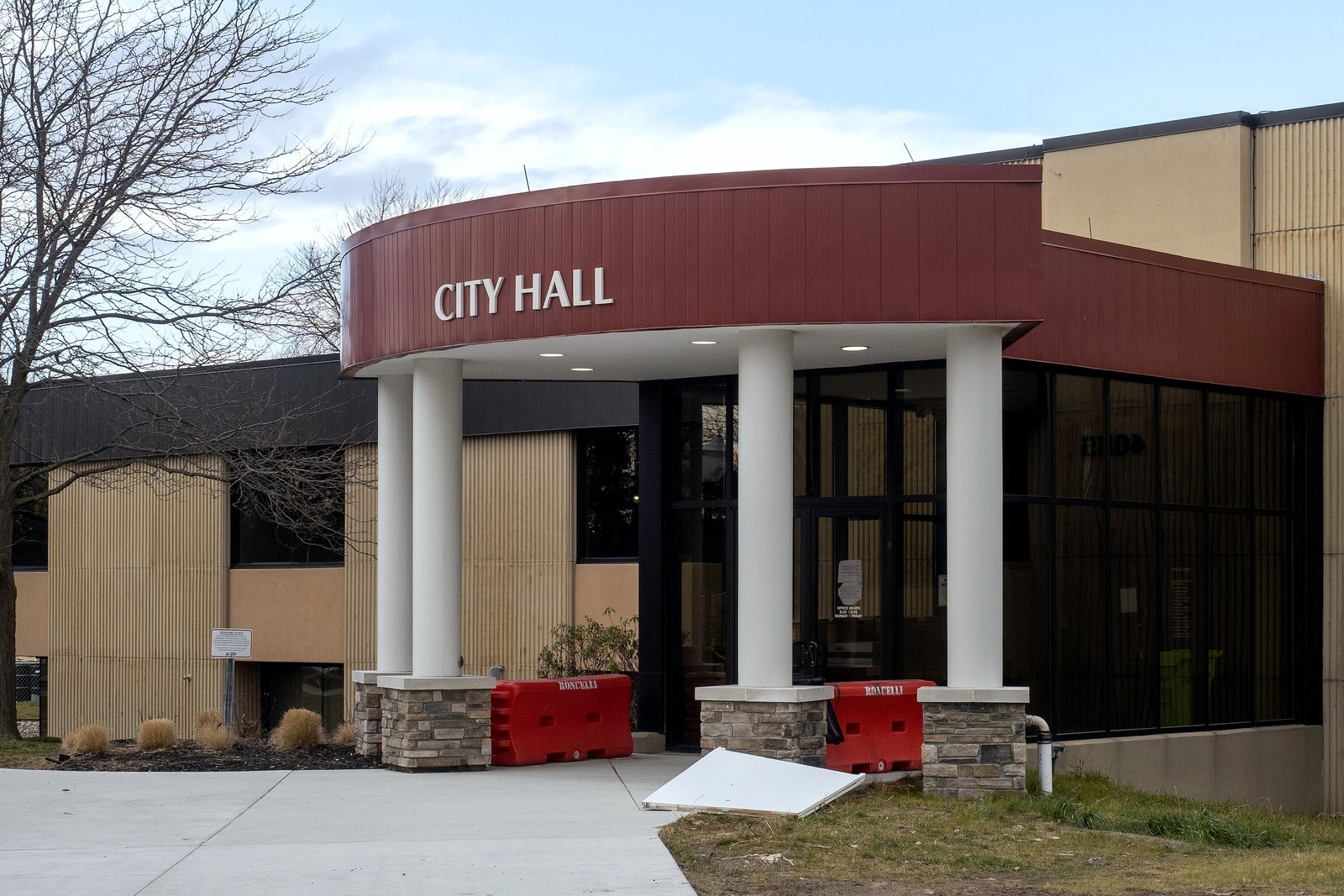 A modern brick and stone city hall with a red-trimmed entrance, columns and a wide plaza with steps in front.