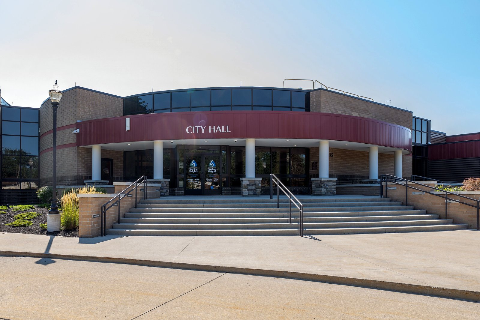 A one-story tan brick building with a curved maroon band reading City Hall above white columns and a wide set of concrete steps.