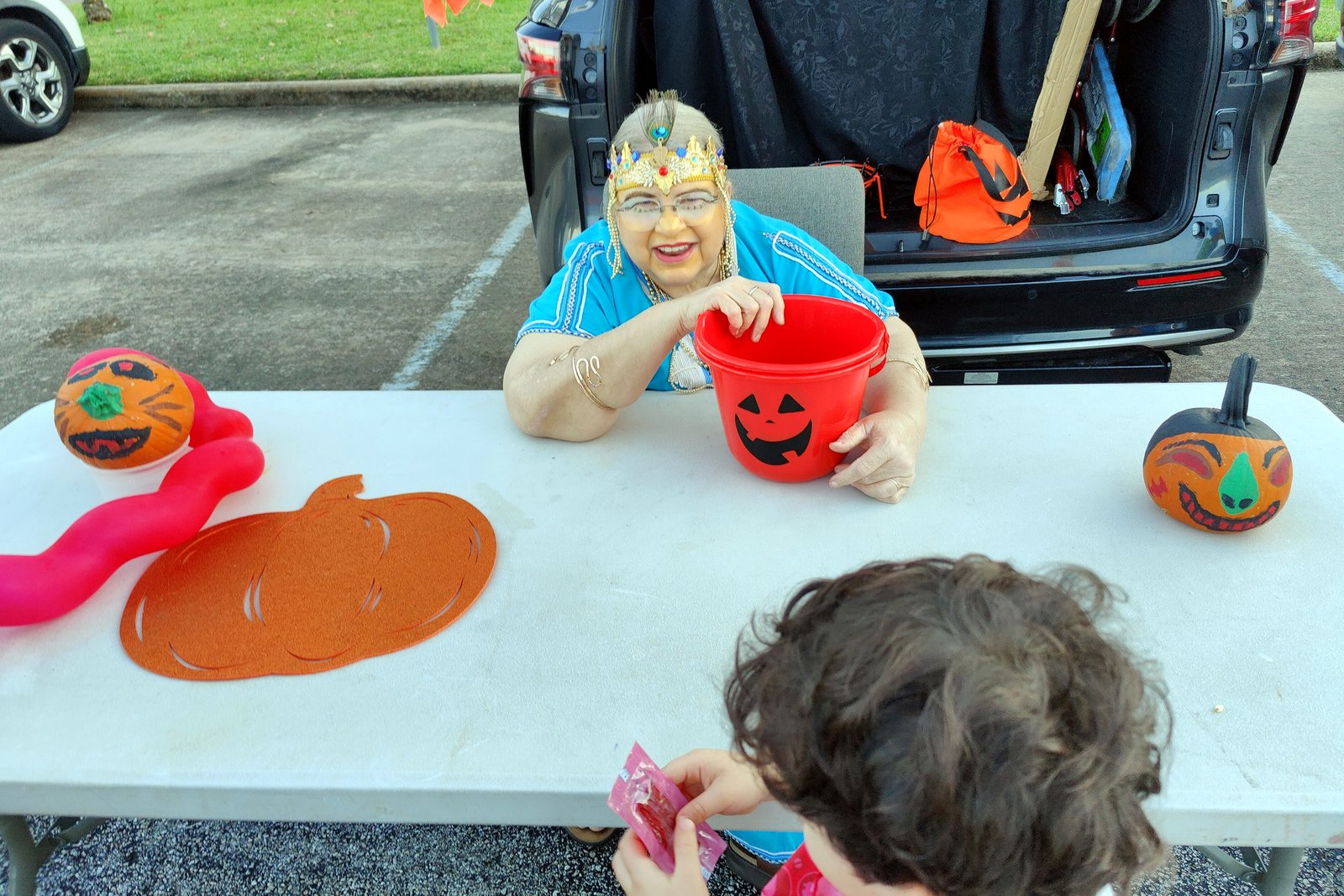 A person in a costume sits behind a table with a pumpkin bucket and decorations at the open back of a car, while a child reaches for candy.