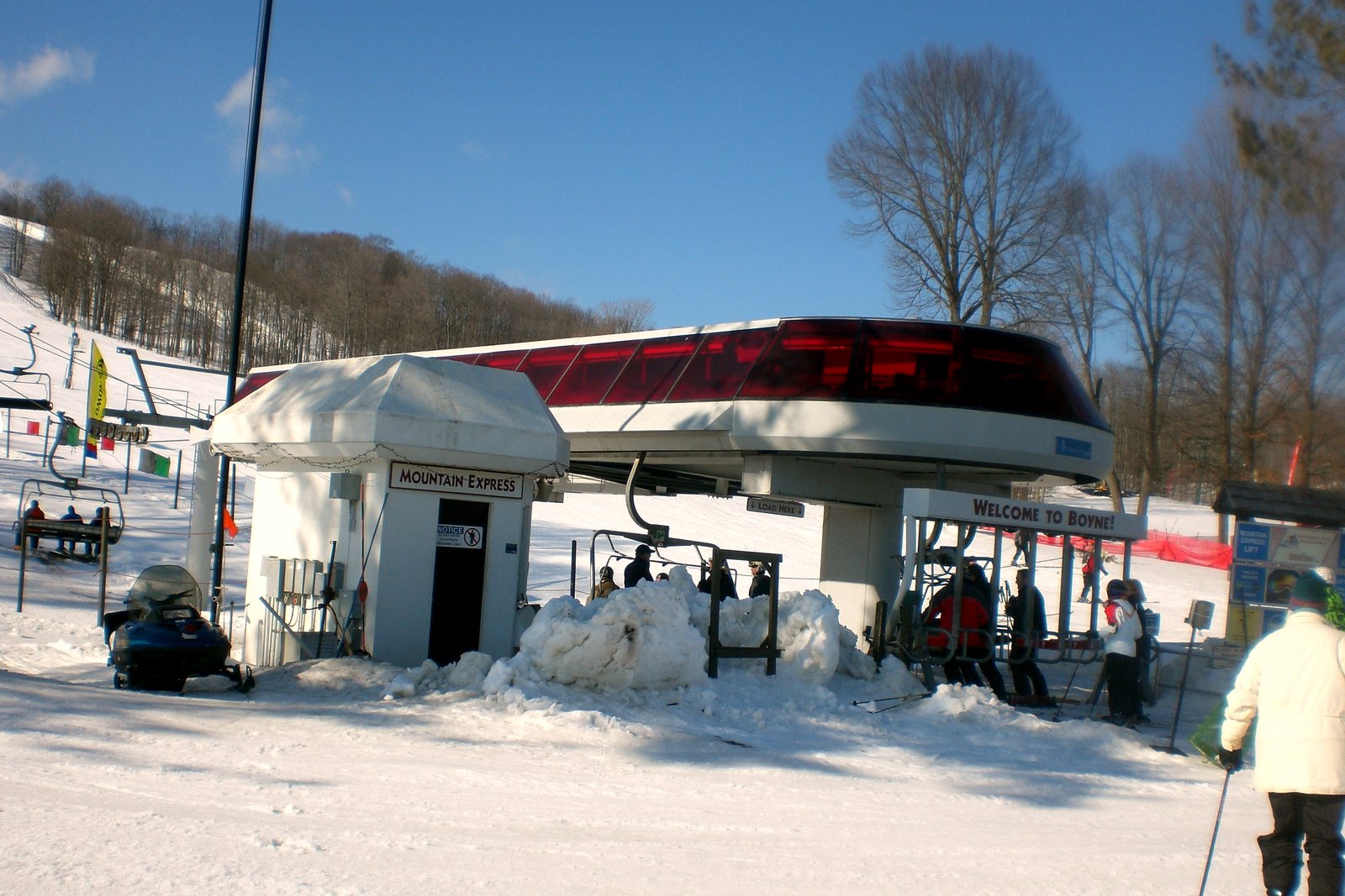 A ski lift terminal with a red canopy and a small white building marked Mountain Express on a snowy slope, with skiers nearby and a wooded hill behind.