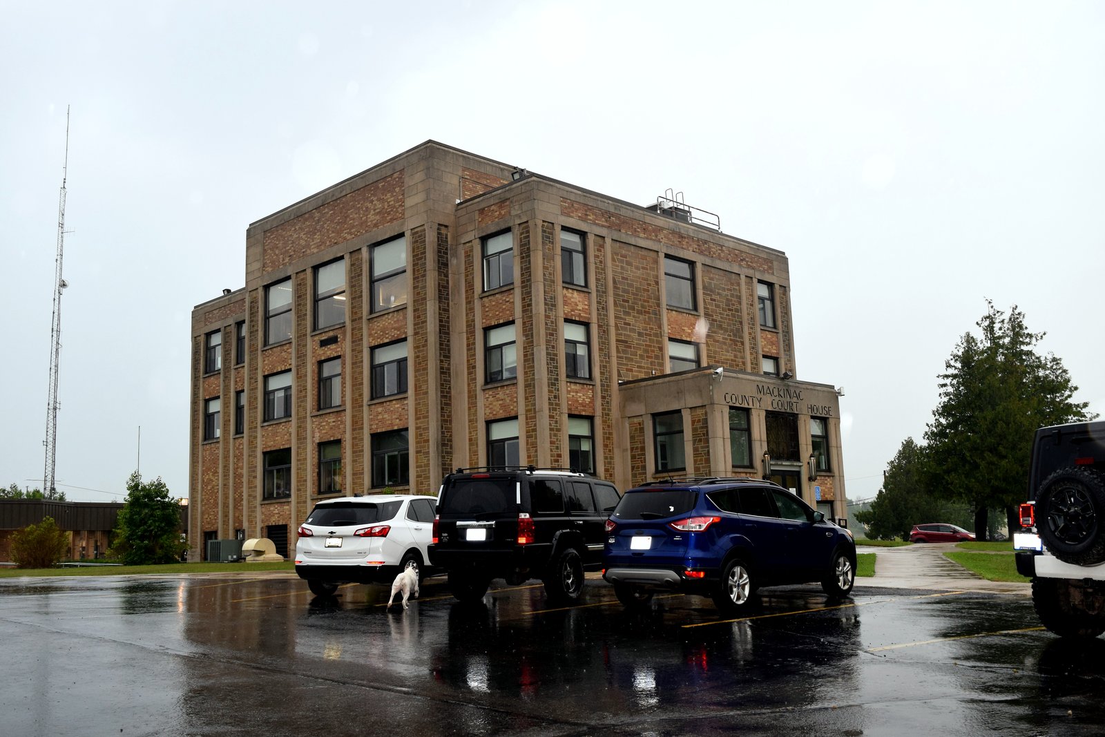 The Mackinac County Courthouse, a three-story tan brick building, stands behind cars parked on wet pavement on a gray day.