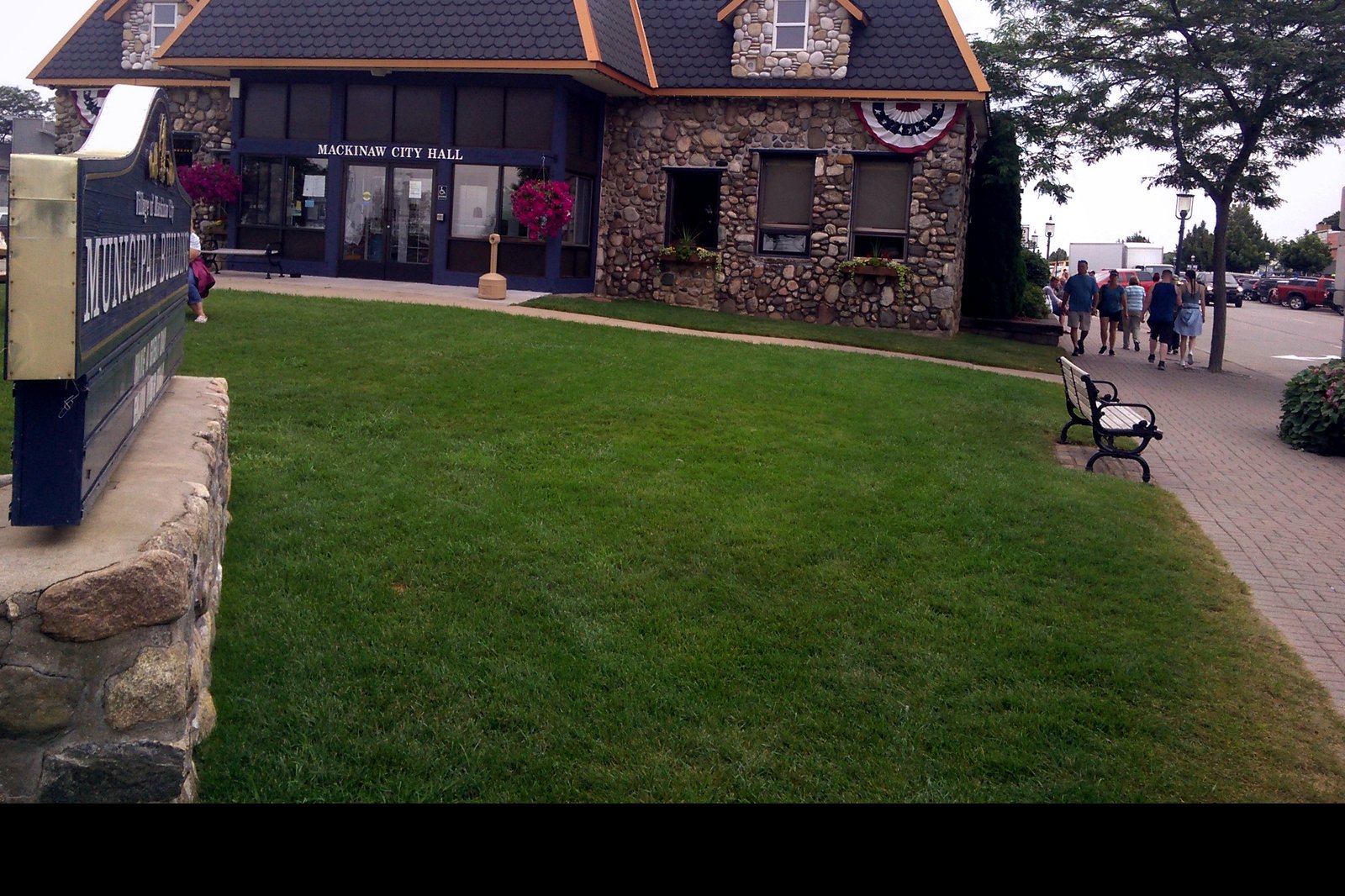 A stone-and-shingle building marked Mackinaw City Hall behind a lawn, with a municipal building sign in the foreground.