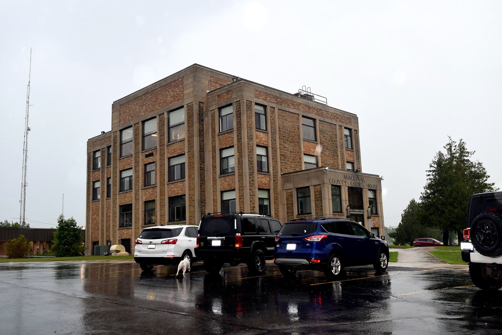 A three-story tan brick courthouse with a flat roof on a rainy day, with several SUVs parked in a wet lot in front.
