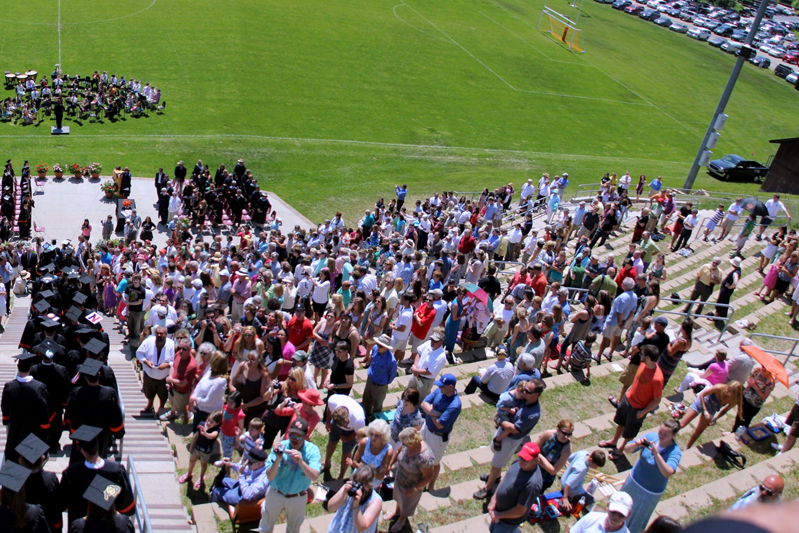 A crowd stands on terraced seating above a green football field with a goalpost during an outdoor graduation ceremony.
