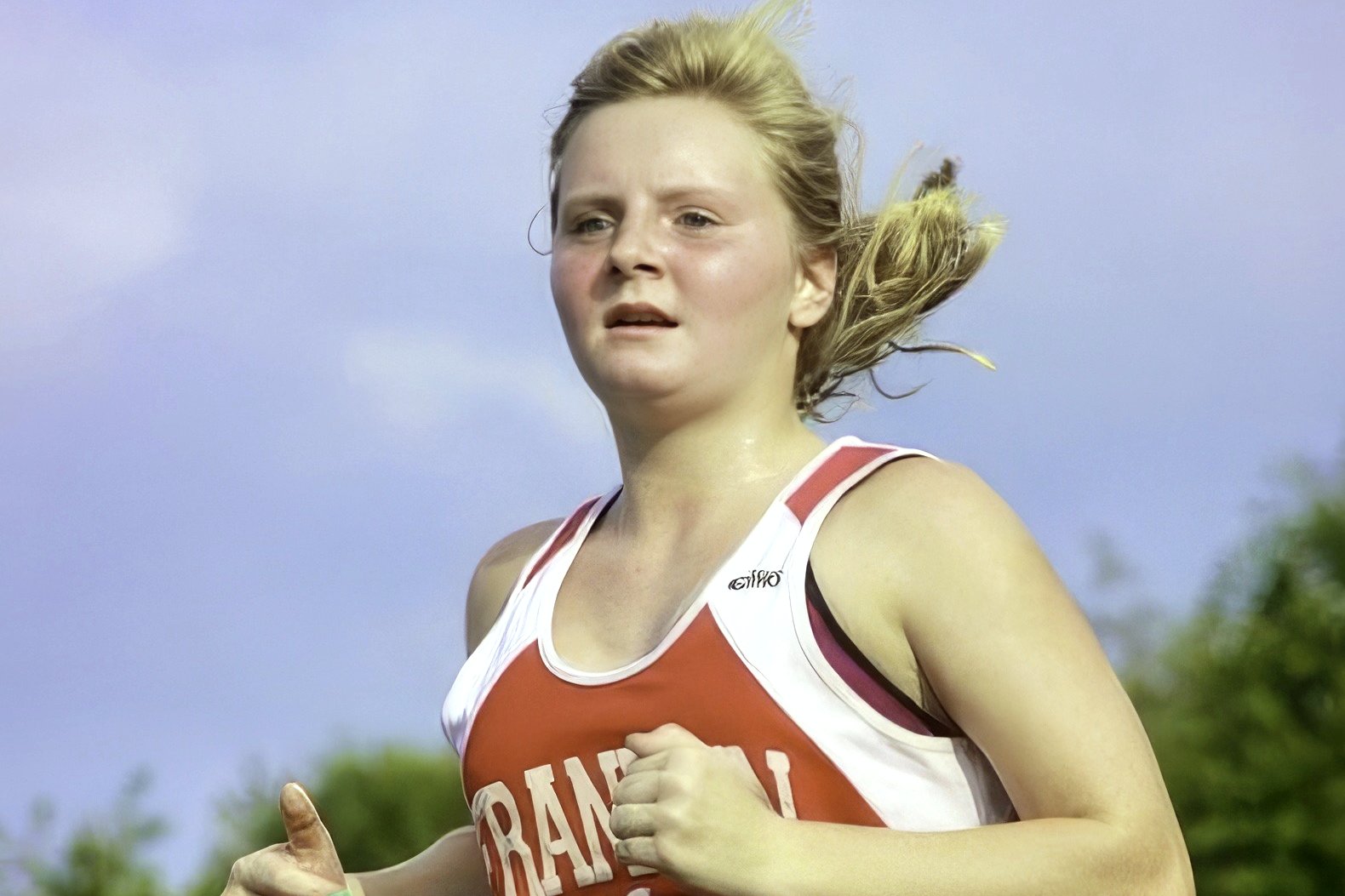 A high school cross country runner in an orange and white singlet runs past green trees under a pale sky.