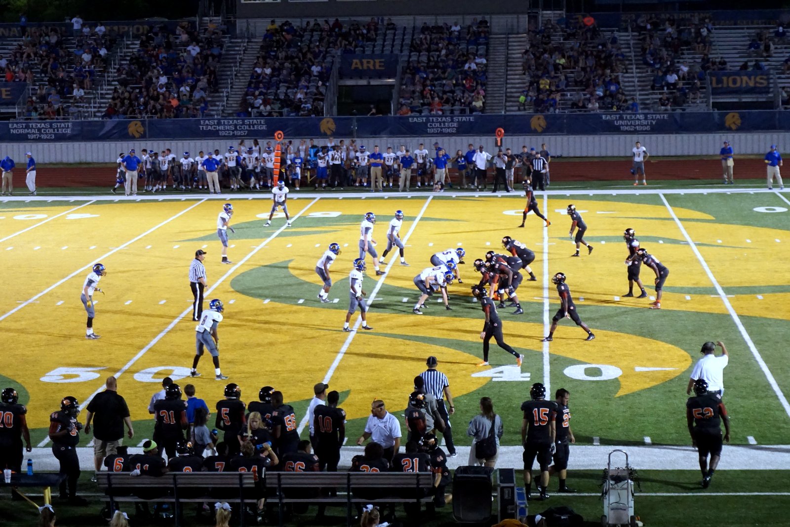 A high school football team runs a play on a green field with a large gold midfield logo as teammates watch from the sideline.