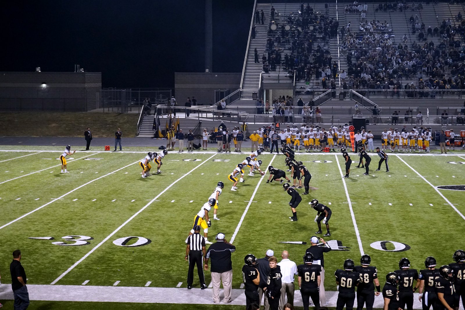 Two high school football teams line up for a punt on a green turf field, with players and coaches watching from the near sideline.