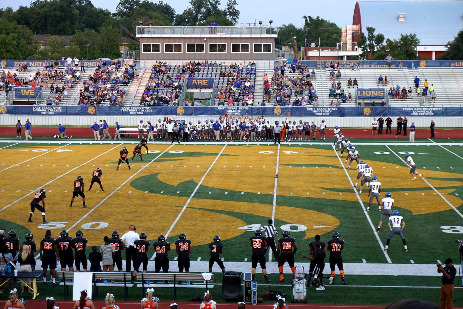 The opening kickoff of a high school football game, seen from behind one team’s sideline, with a crowd in the stands across the field.