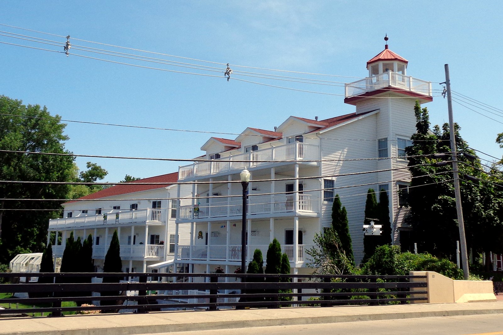 A white multi-story building topped with a small lighthouse tower and red roof stands near the water in Caseville, with trees and a road in front under a blue sky.