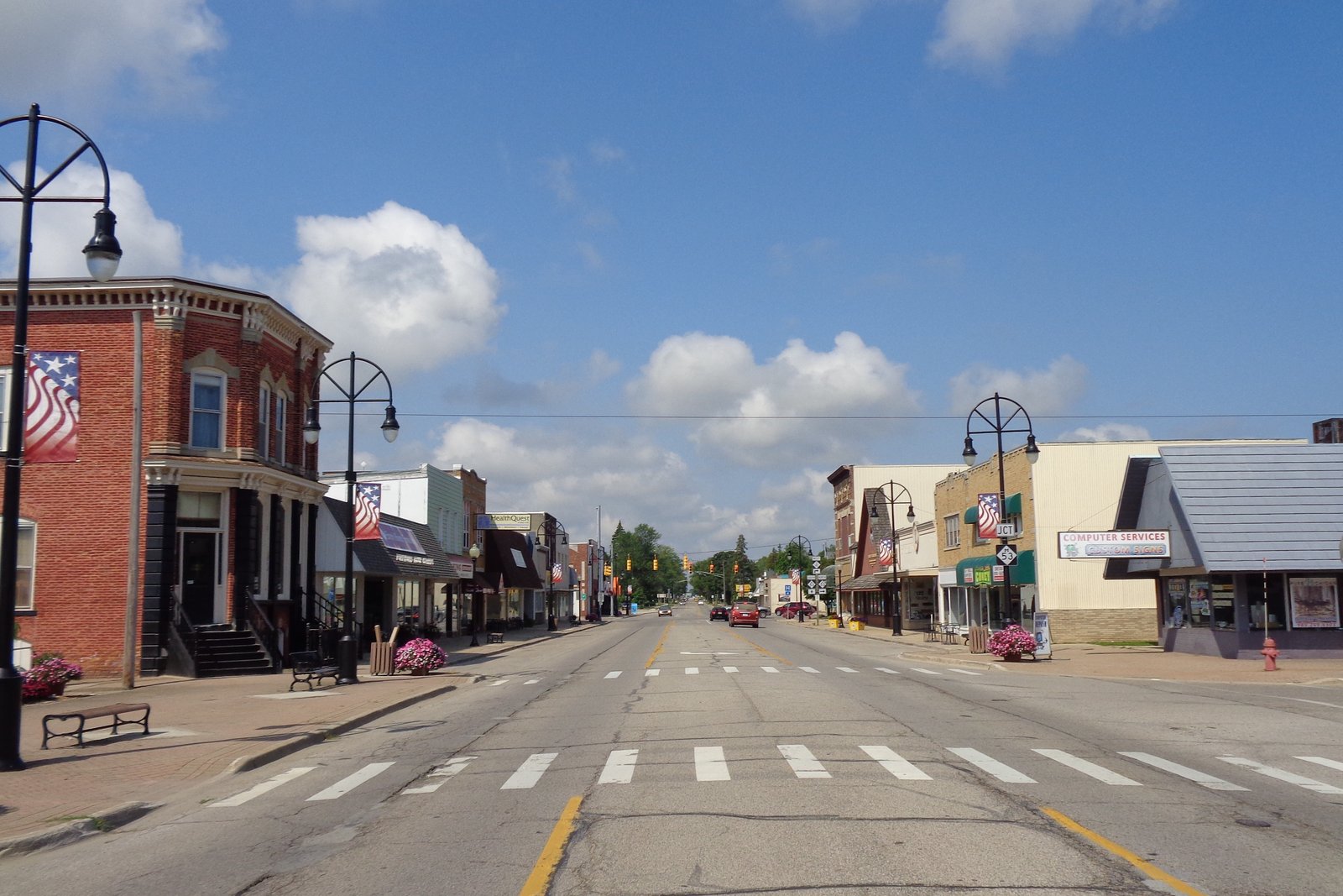 Downtown Bad Axe along M-142, with brick storefronts, black lampposts, flower planters and a crosswalk under a blue sky.