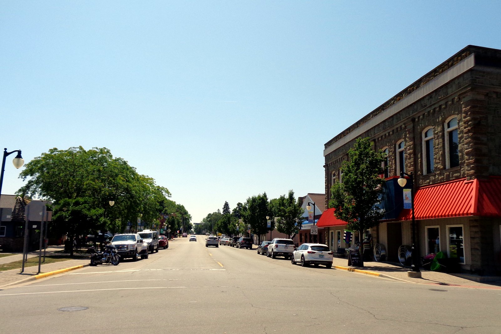 A wide main street in downtown Port Austin on a sunny summer day, with a stone commercial building under red awnings on the right and cars parked along the road.