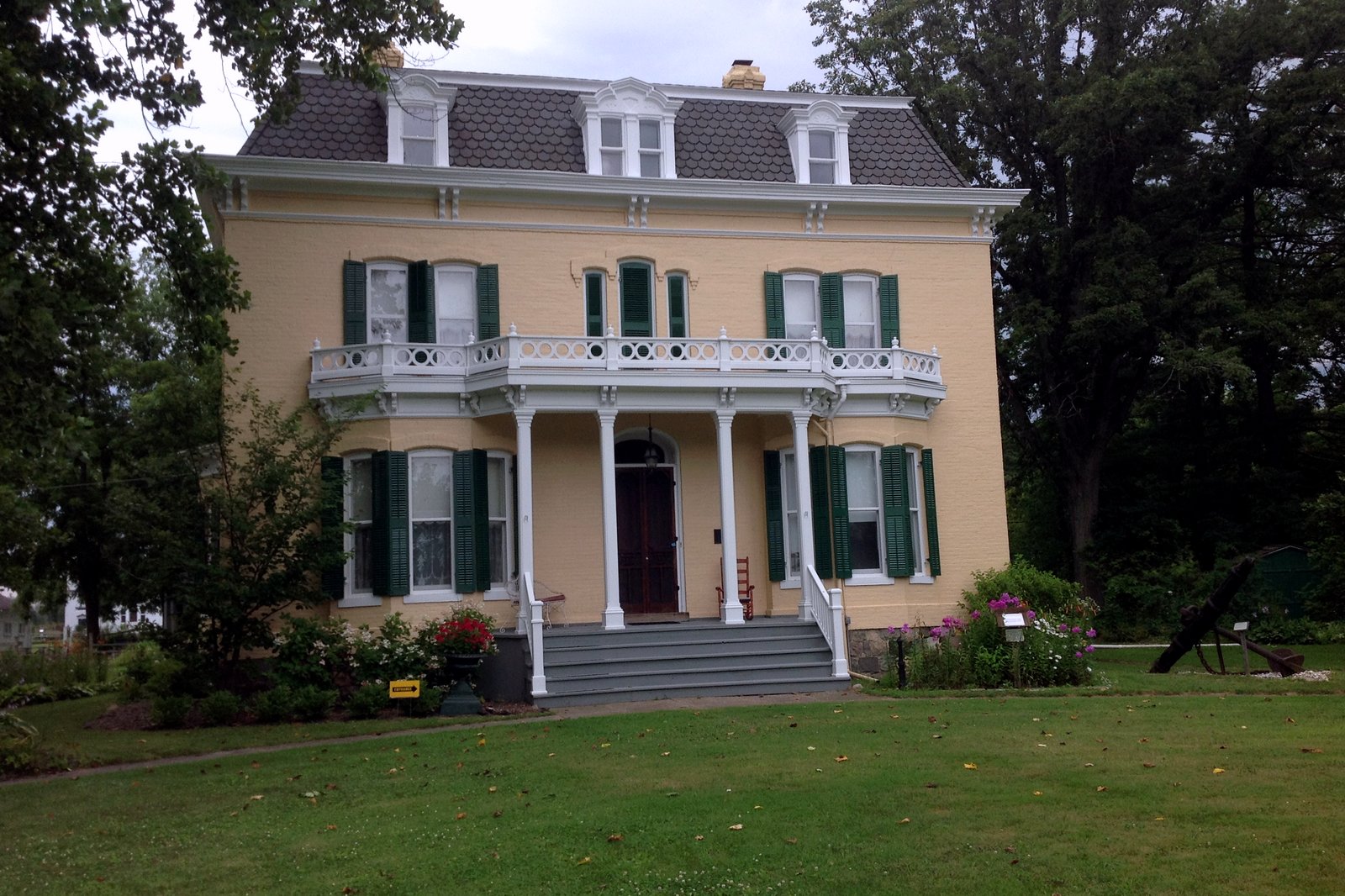 A two-story cream brick house with green shutters, a white columned porch and a balcony, set behind a lawn and flower beds and framed by tall trees.