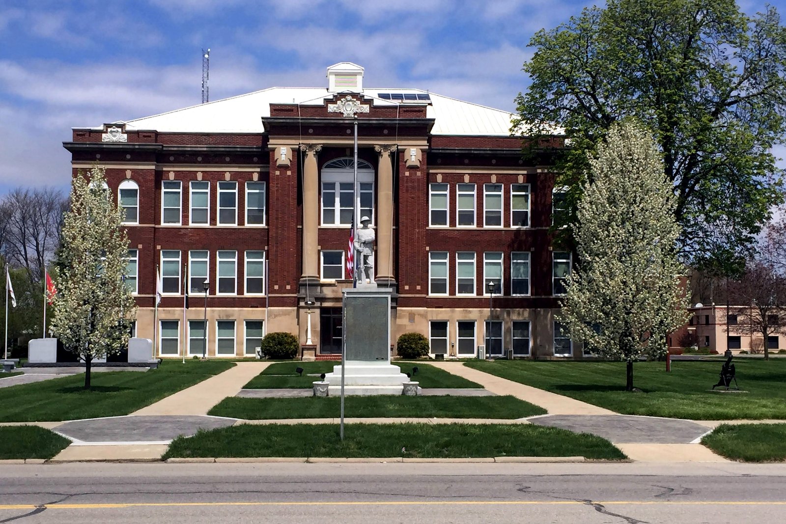 A three-story red brick courthouse with white trim, a soldiers’ monument and flagpole on the front walk, and flowering trees on the lawn.