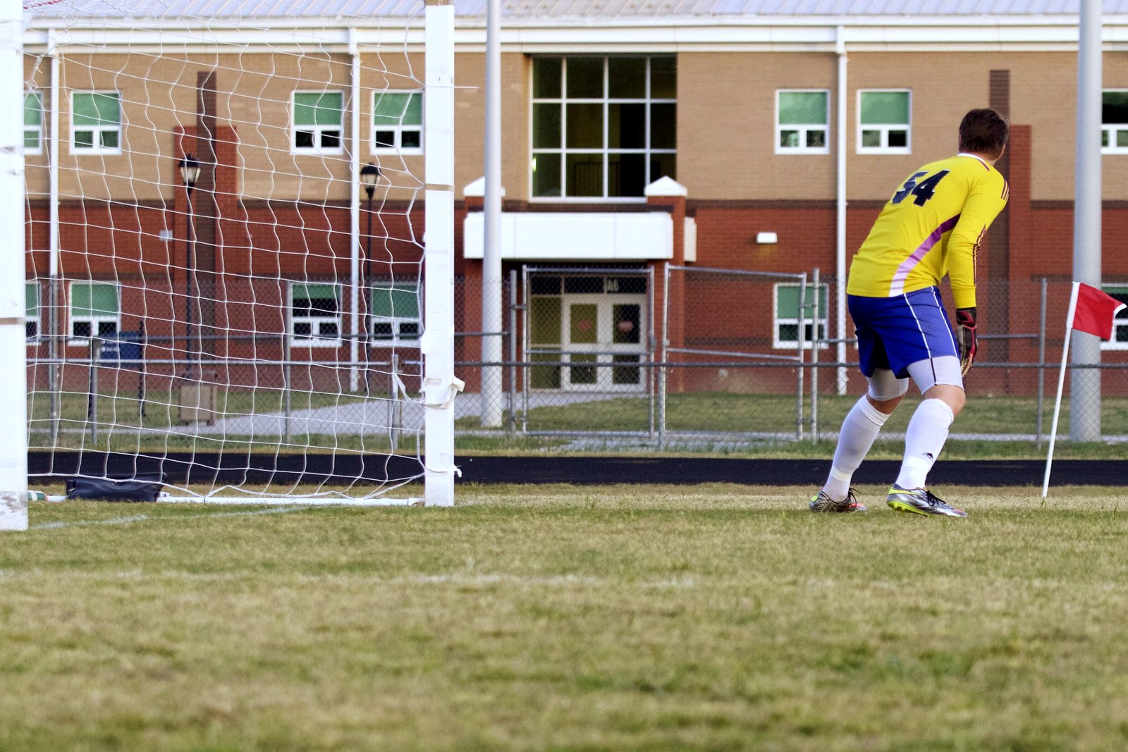 A soccer goalkeeper in a yellow jersey and blue shorts stands in front of the goal net on a grass field, with a brick school building behind.