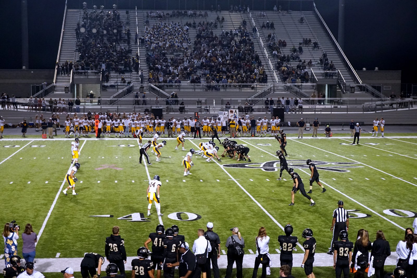 Football players in white and gold uniforms line up on offense against a team in black on a lit field, with full stands and a sideline of players behind them.