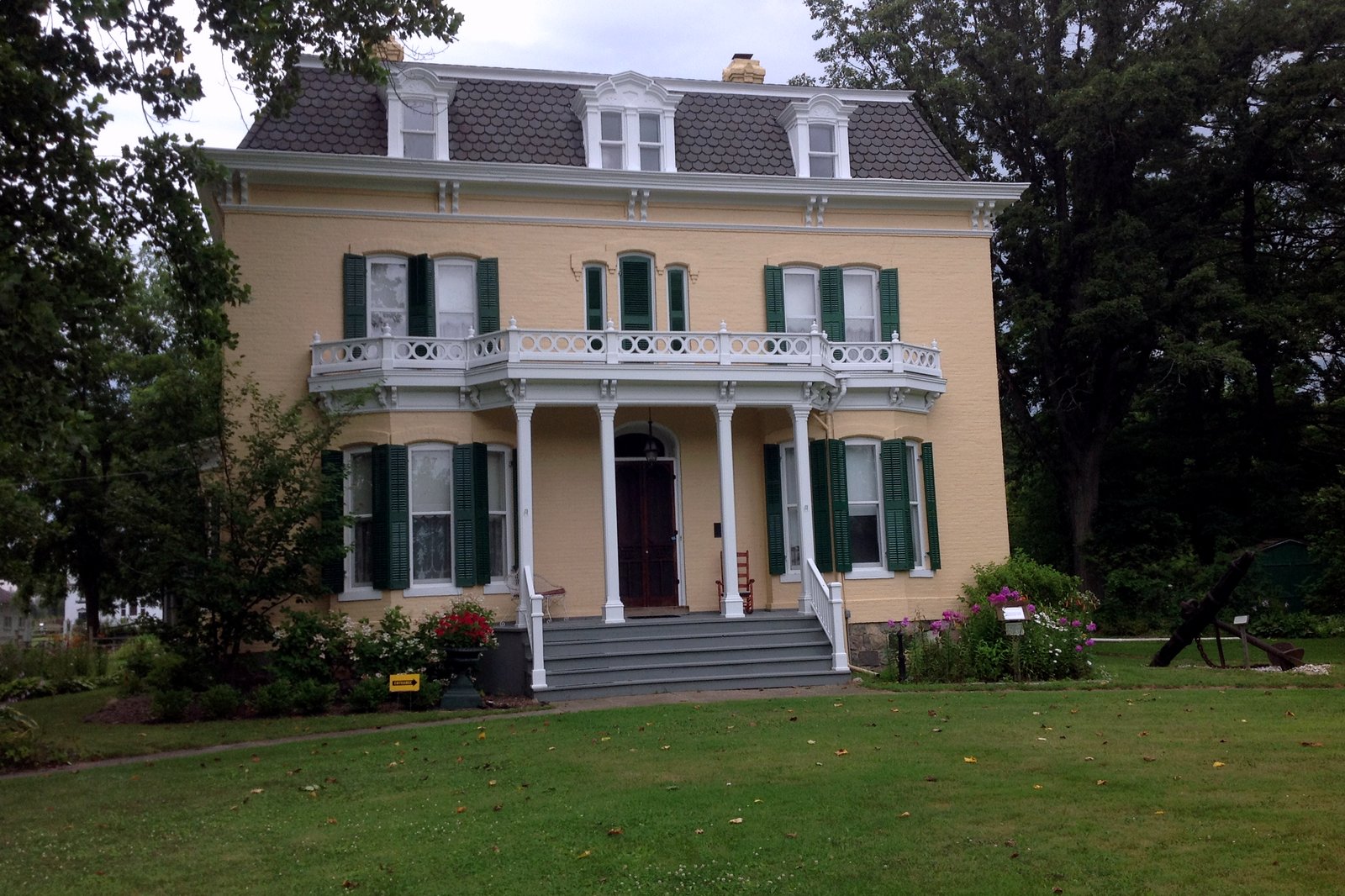 A two-story cream-colored brick mansion with green shutters, a white columned porch with a balcony and a mansard roof, set on a green lawn among trees.