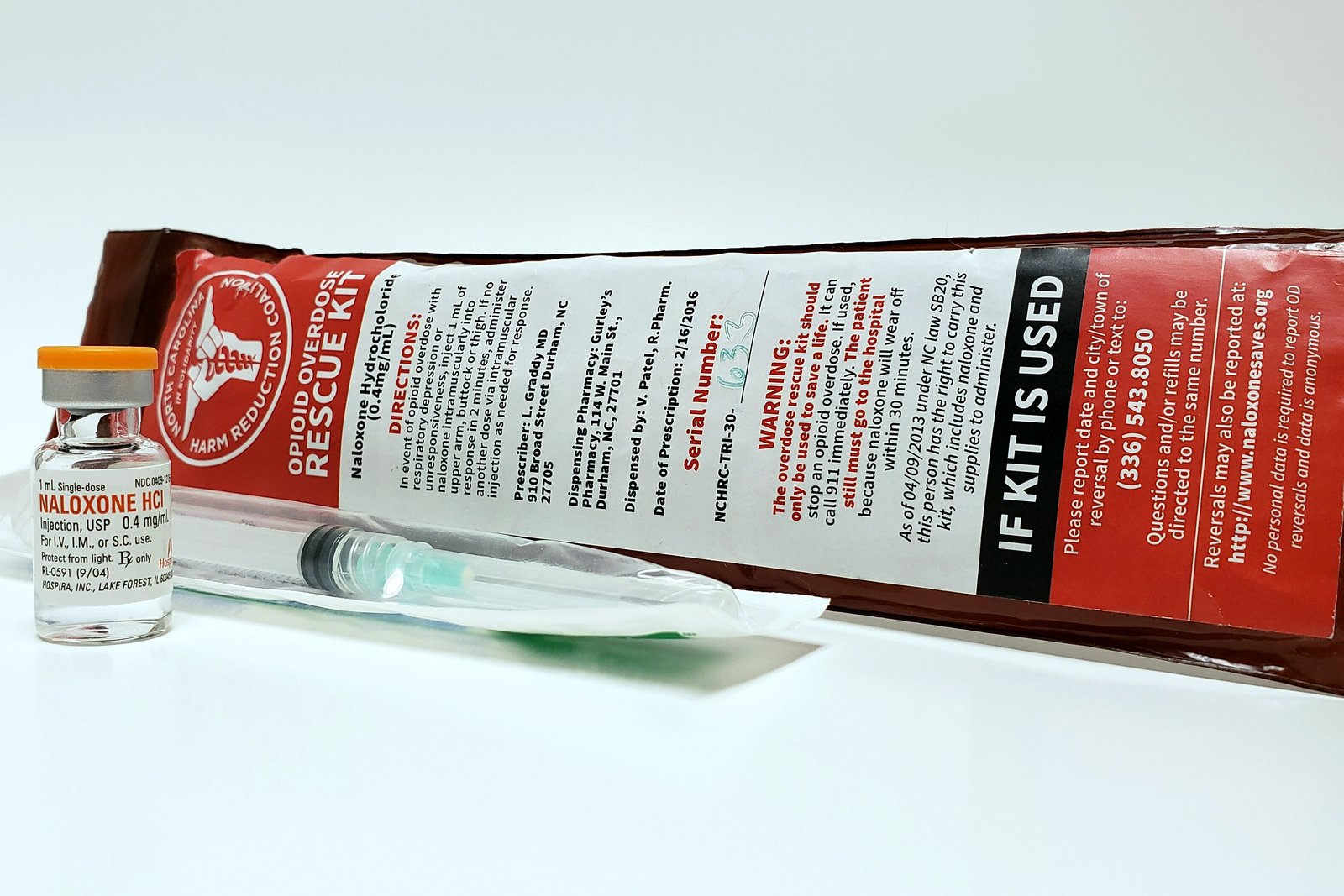 A small vial of naloxone and a syringe lie in front of a red and white opioid rescue kit package on a white surface.