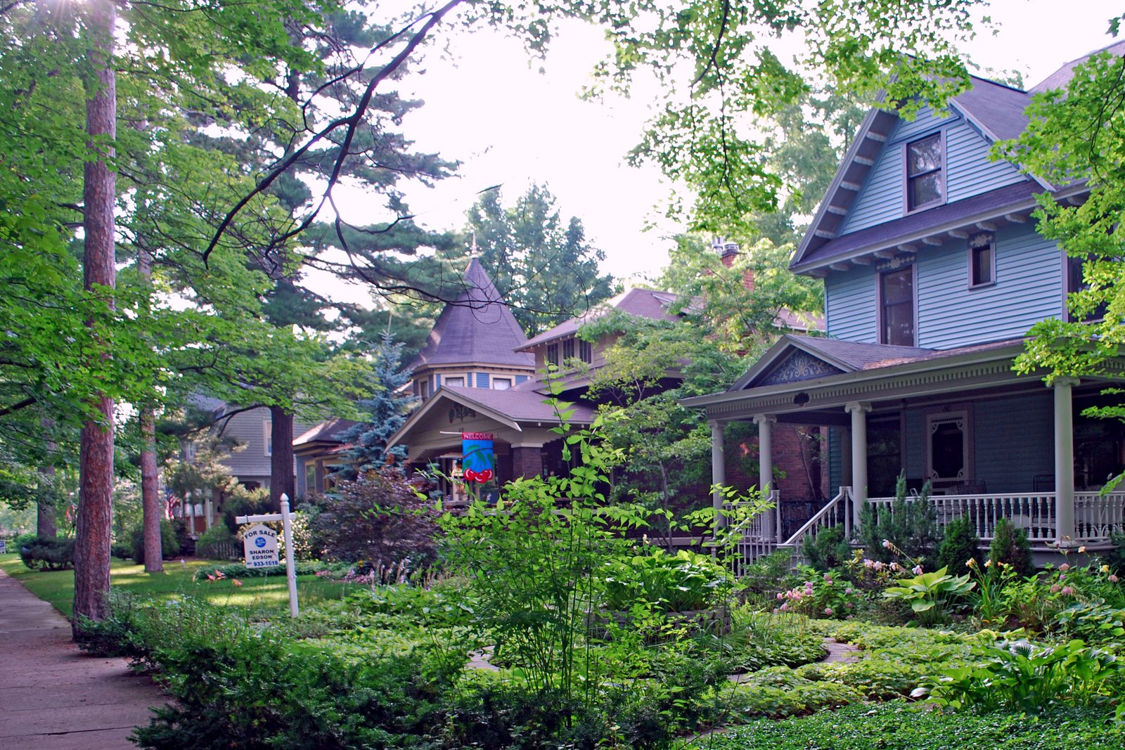 Large older houses with porches and a turret among tall trees and garden beds along a sidewalk on a summer day.