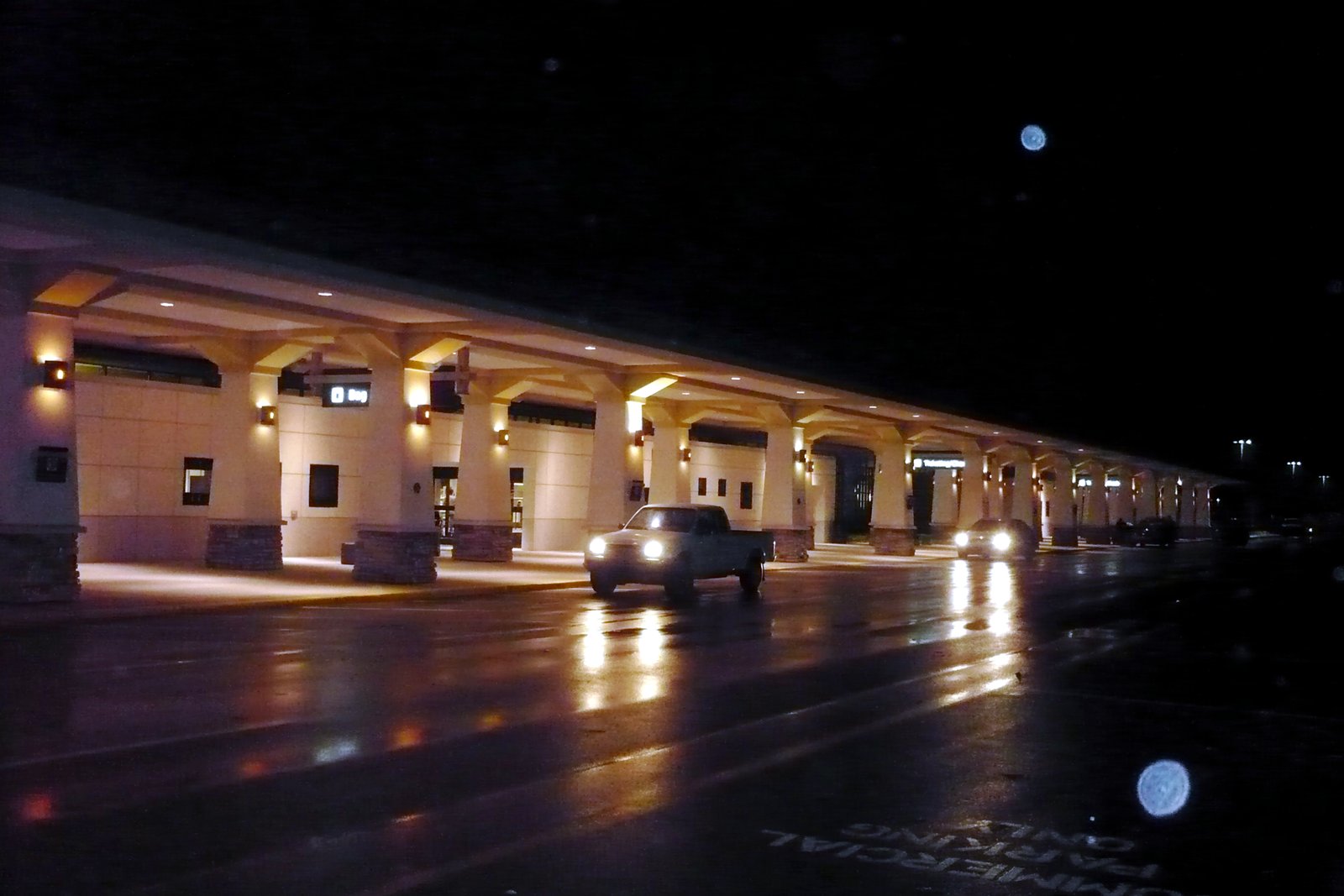 A covered airport terminal curb at night, lit by lamps, with a pickup truck driving along the wet road in front.