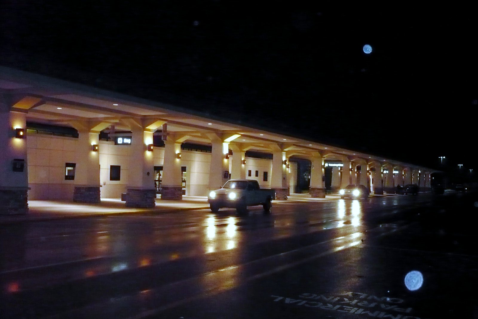 An airport terminal’s covered curbside lane lit up at night, with a car passing on the wet pavement.