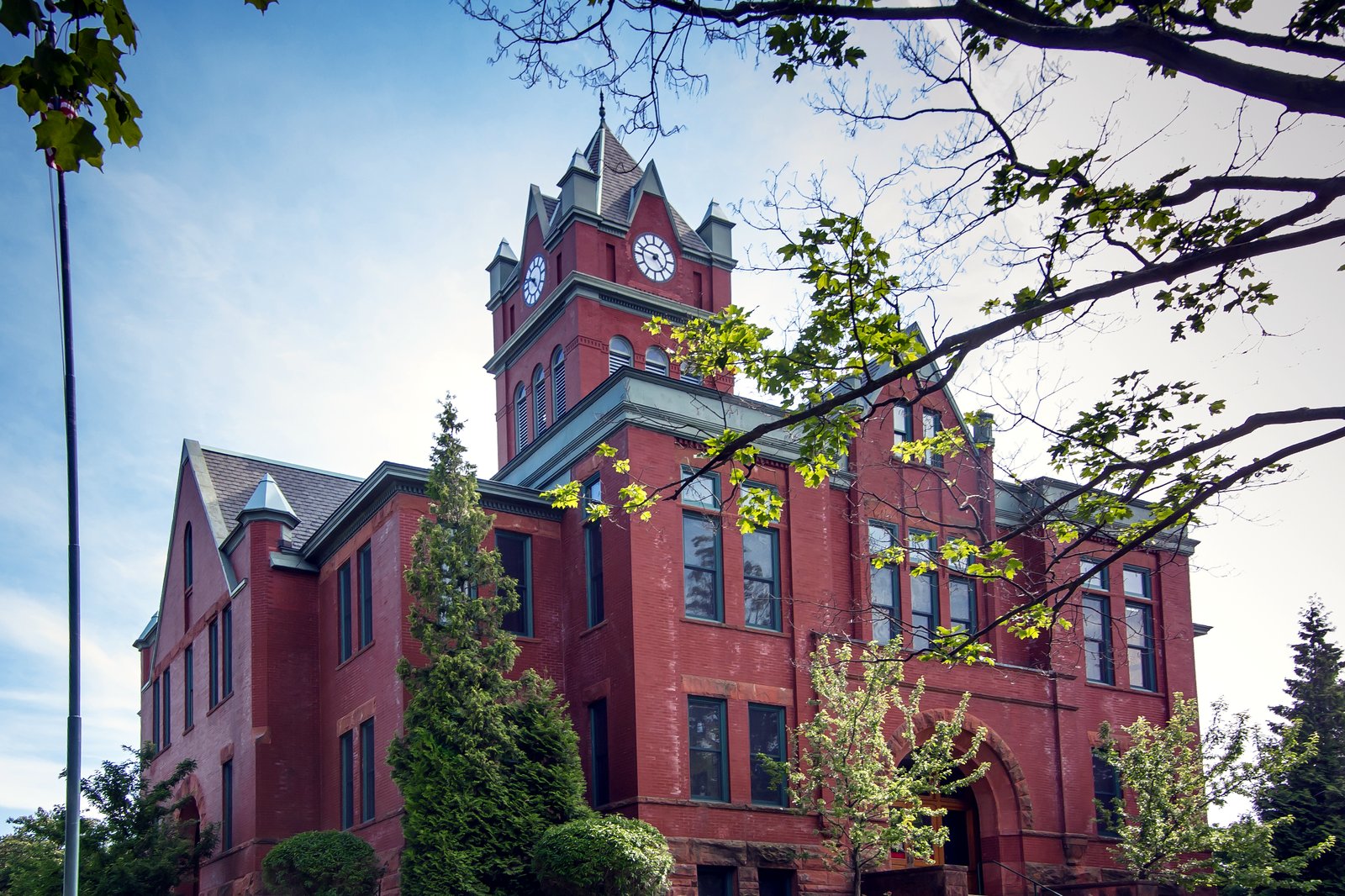 A red brick courthouse with a tall clock tower and arched windows stands on a grassy rise behind trees under a blue sky.