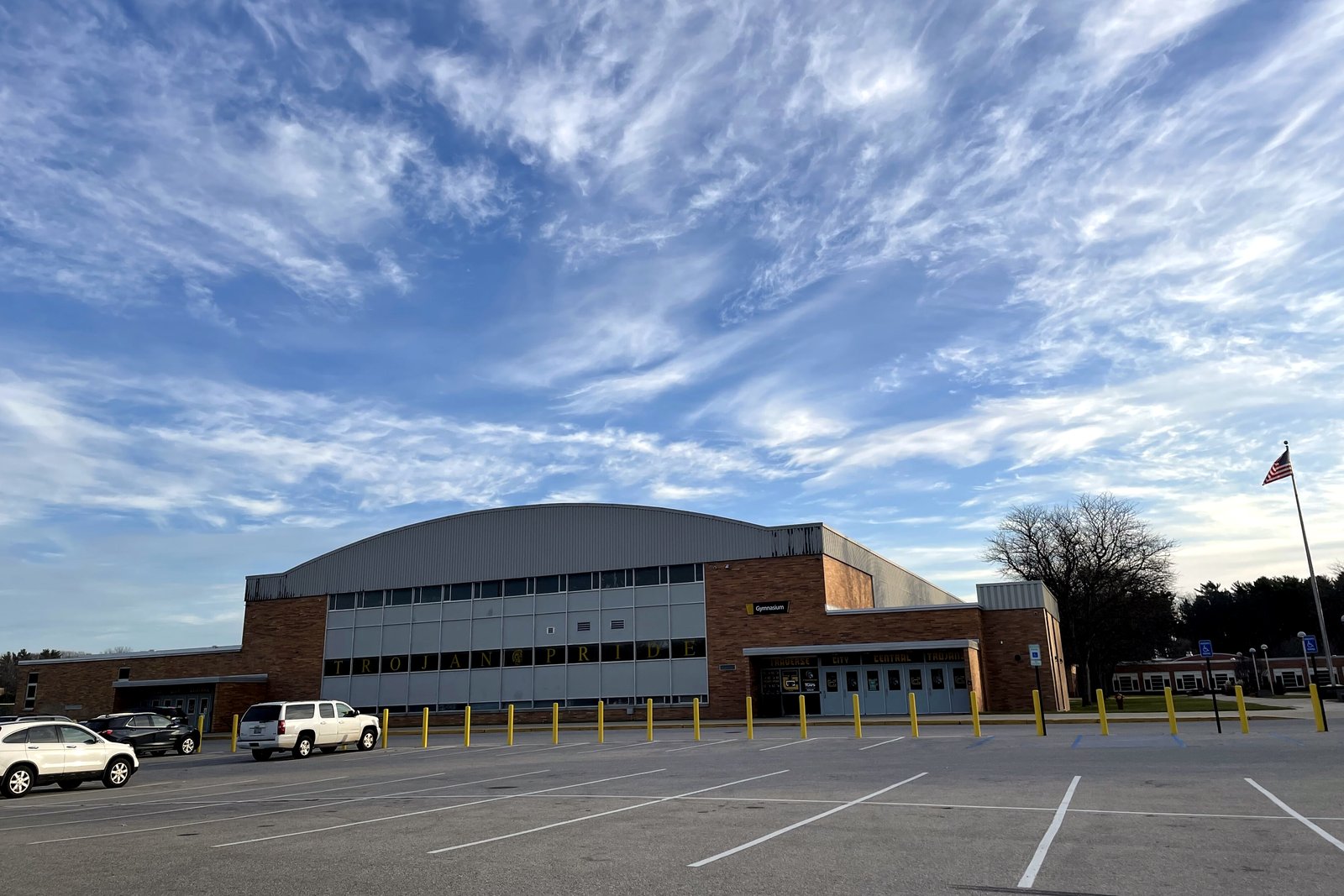 A long low brick school building with a rounded gym roof behind a parking lot, under a sky of wispy clouds.