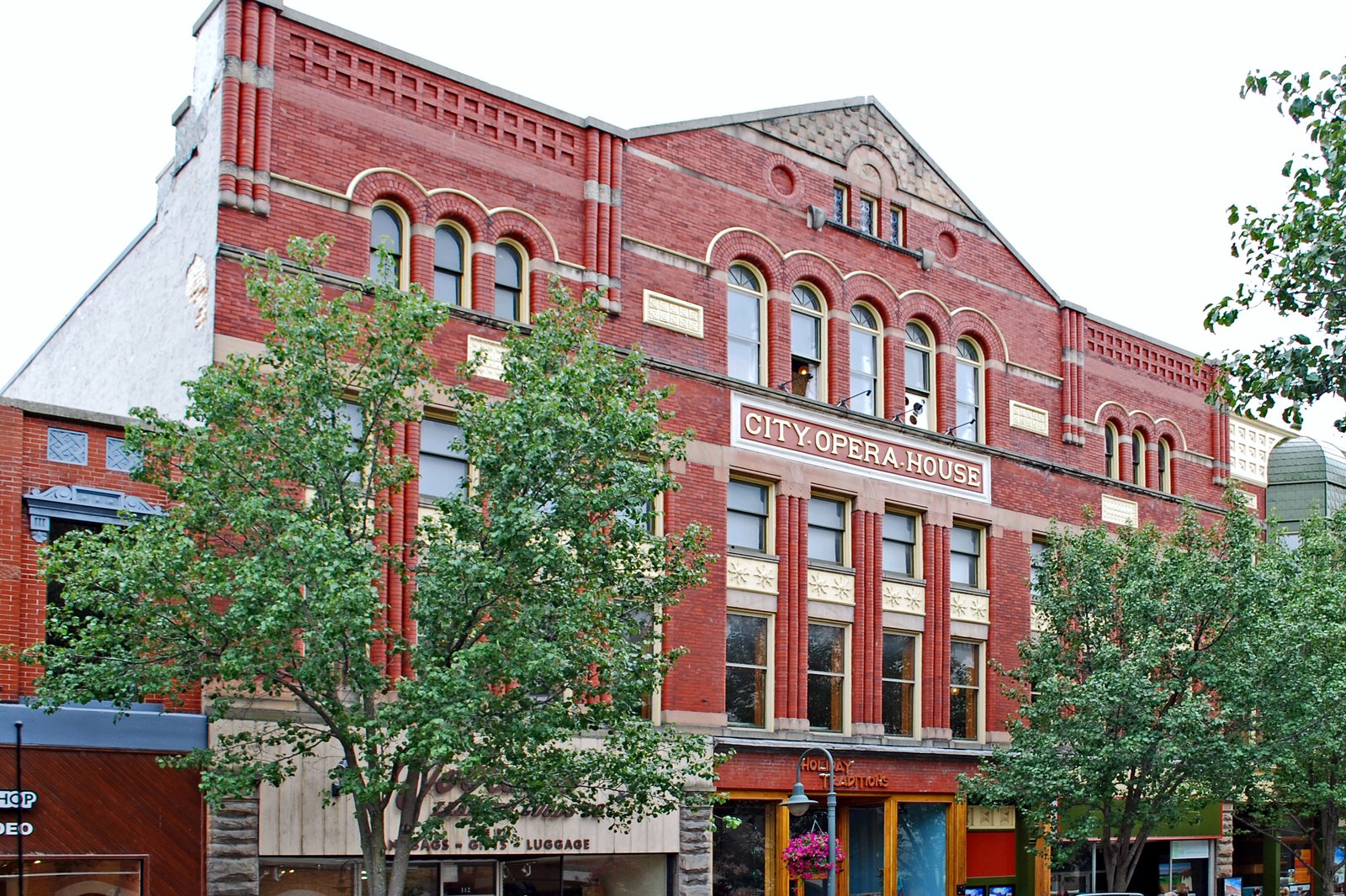 A red-brick Victorian commercial building with tall arched windows and a decorative cornice, with trees and a parked car in front.