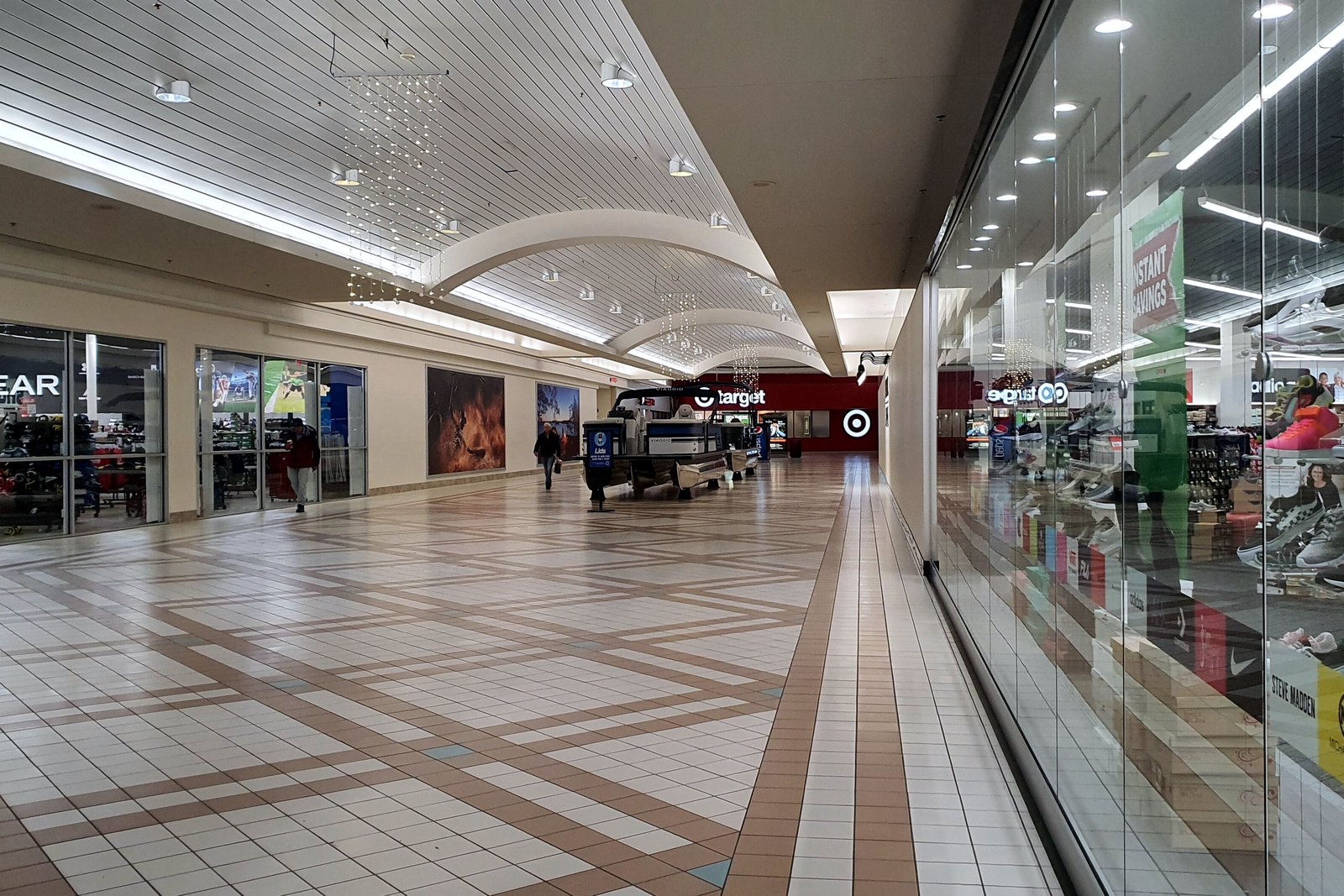 A wide, bright shopping mall concourse with a tiled floor, skylights, store entrances and a few shoppers.