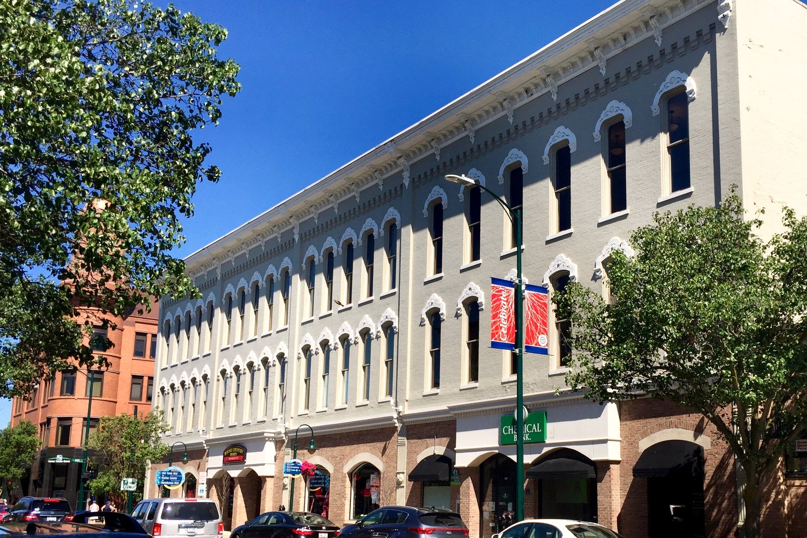 A long three-story downtown block with rows of arched windows, street trees and banners on light poles under a blue sky.