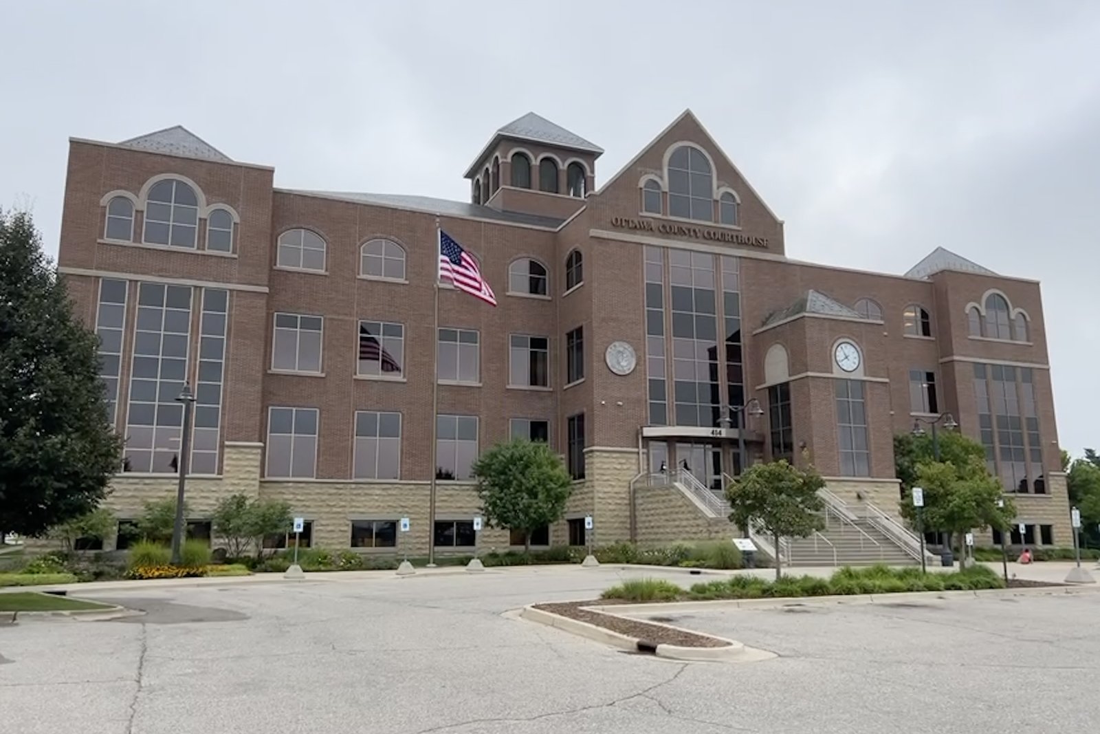 The four-story brick Ottawa County Courthouse in Grand Haven, with arched windows, a cupola and an American flag out front, under a cloudy sky.