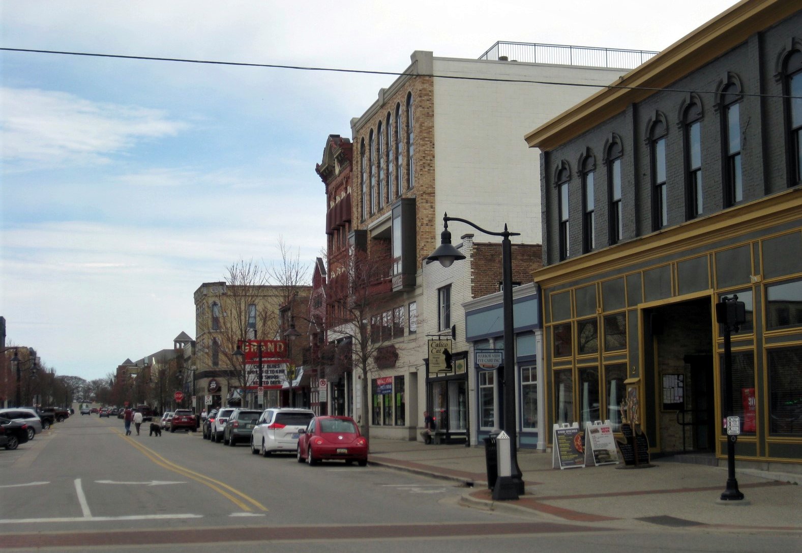 Brick and stone storefront buildings line a downtown Grand Haven street, with the Grand Theatre building on the right and cars parked along the curb.