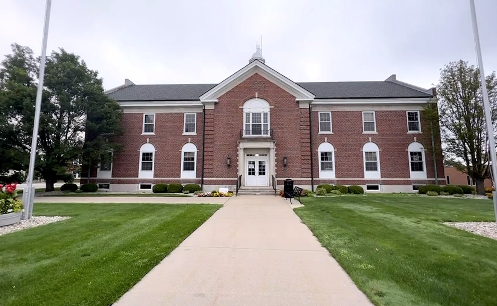 A two-story red-brick building with white-trimmed arched windows, a central gable and a small cupola, set behind a wide lawn and a concrete walk between two flagpoles under a gray sky.