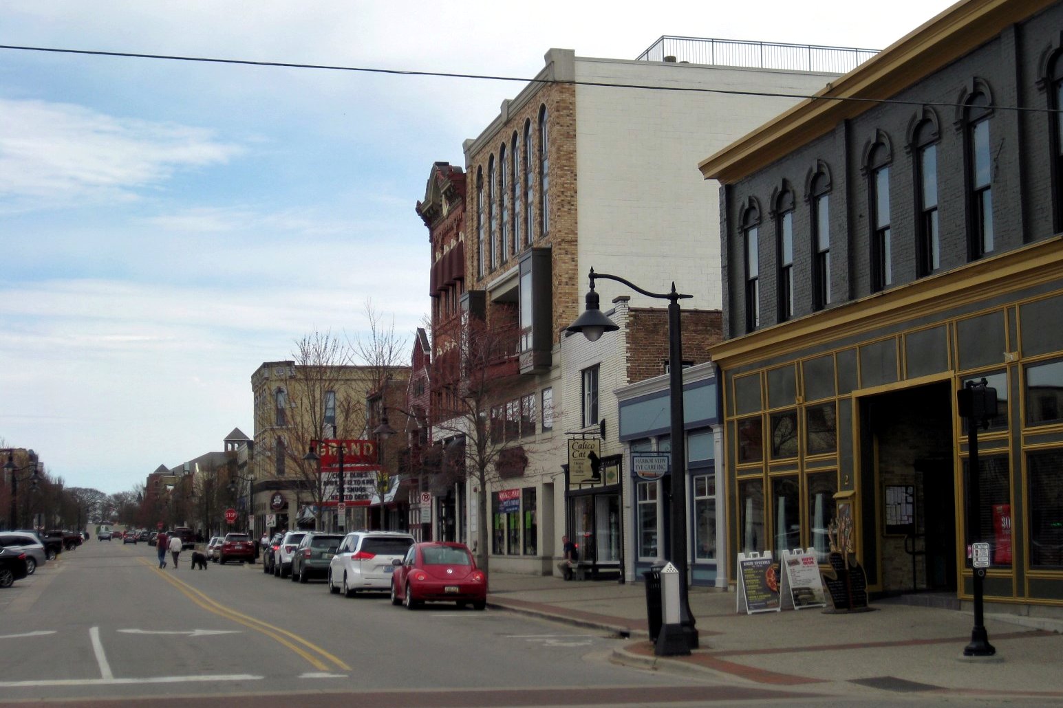 A downtown street of two- and three-story brick and stone storefronts with parked cars and a few pedestrians under a partly cloudy sky.