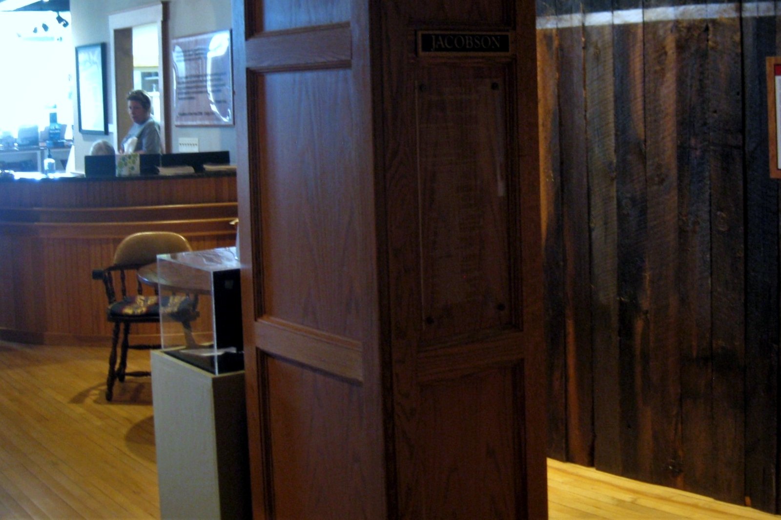 Inside the Tri-Cities Historical Museum in Grand Haven, a wood-paneled front desk and a carved wooden post stand beside a weathered barn-wood exhibit wall on a light wood floor.
