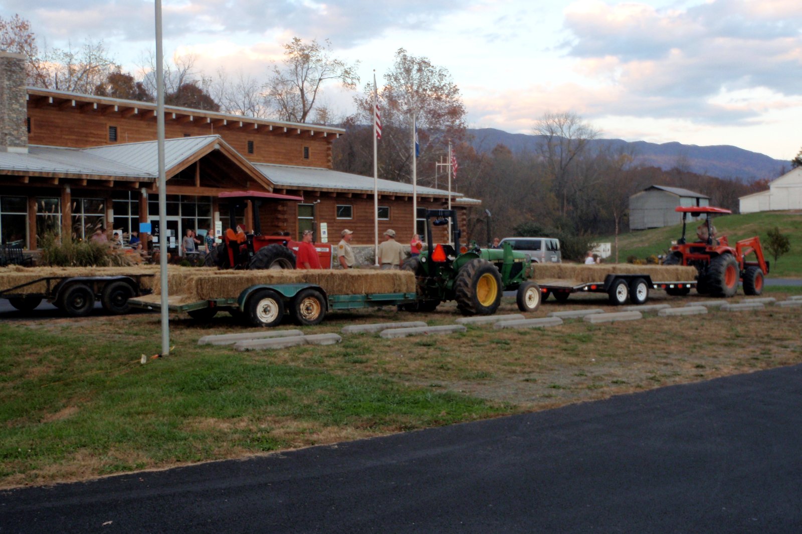 Hay wagons hitched to tractors wait outside a wooden lodge at dusk, with wooded hills behind.