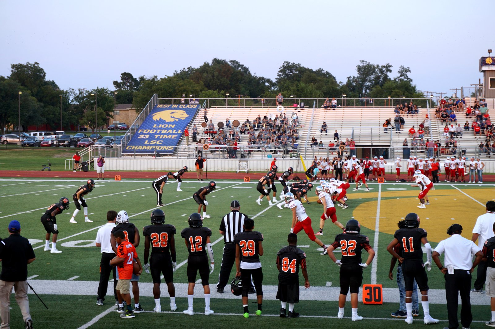 High school football players in dark jerseys line up on offense on a lighted field as defenders wait across the line of scrimmage.