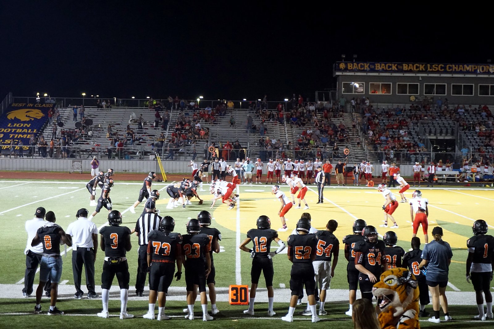 High school football players run a play on a lit field at night as a team in dark orange jerseys watches from the sideline in the foreground.