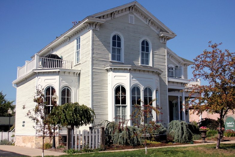 A two-story white clapboard house with arched windows, a bay window and small balconies under a blue sky, with a sign reading Isabel’s House on the lawn.