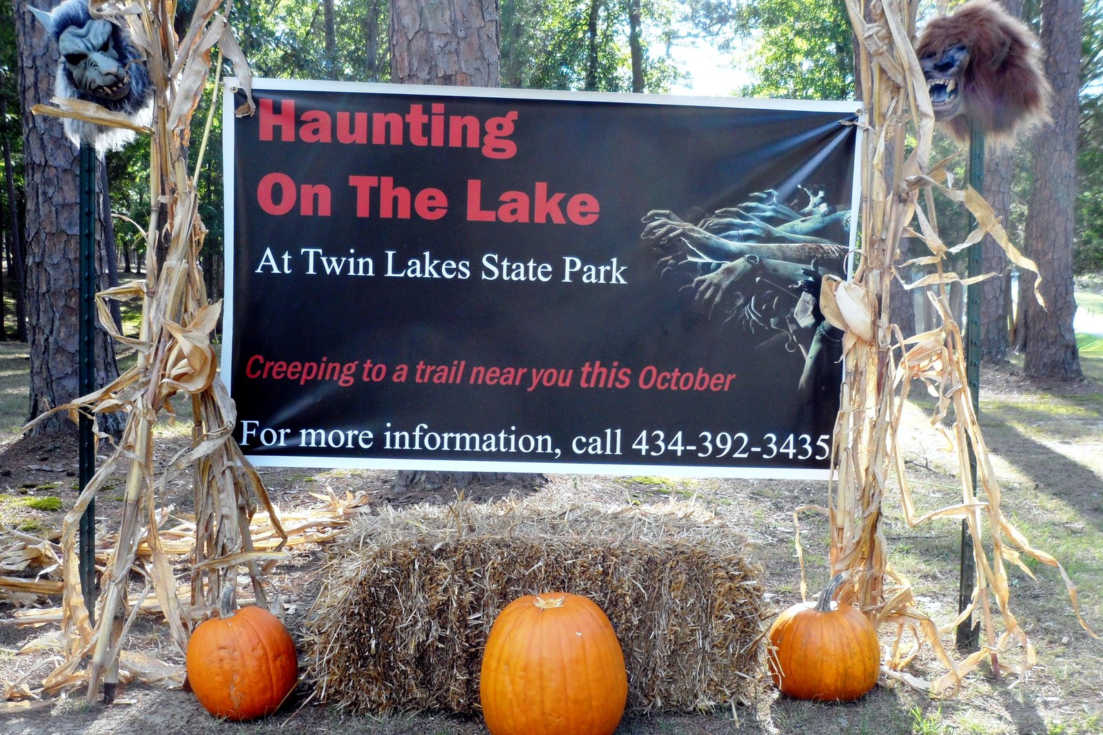 Orange pumpkins and a hay bale sit beneath a banner advertising a Halloween trail event, flanked by corn stalks and Halloween props in a wooded park.