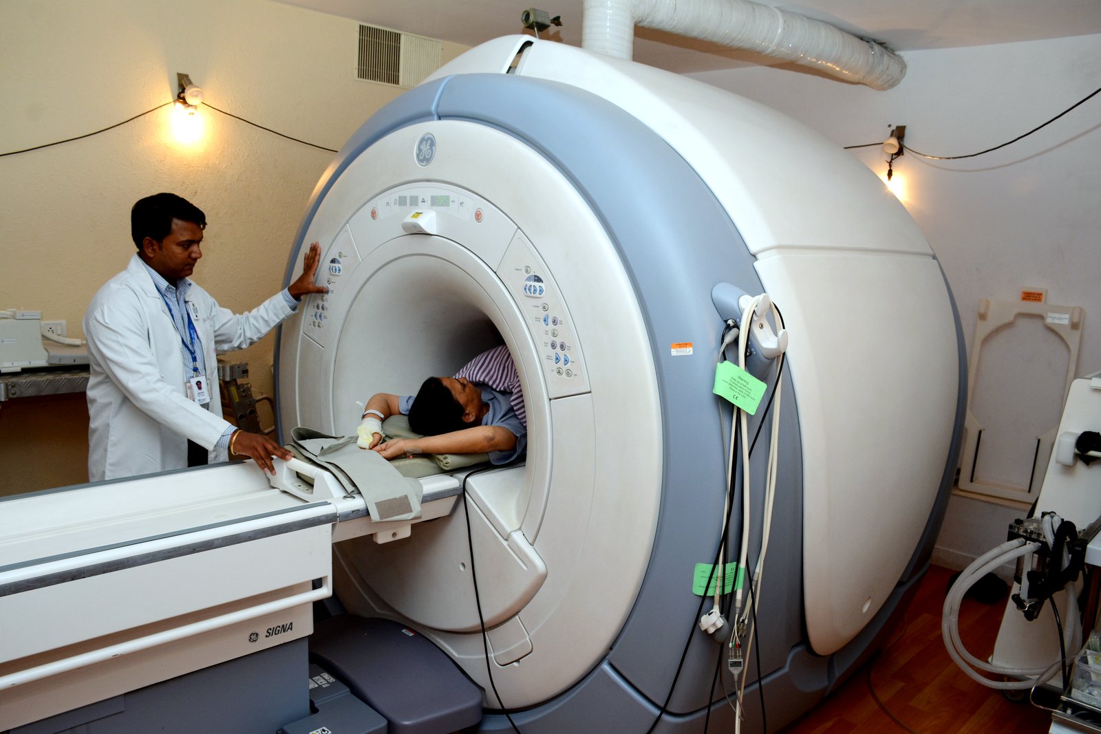 A technician in a white coat stands beside a large MRI scanner as a patient lies on the scanner table.