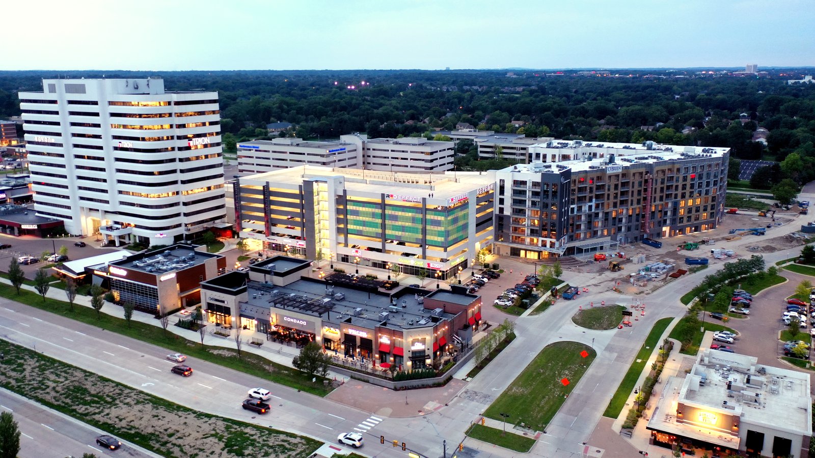 An evening aerial view of a cluster of new mid-rise apartment and office buildings beside a wide divided road, with green lawns, parking lots and a flat tree canopy stretching to the horizon.