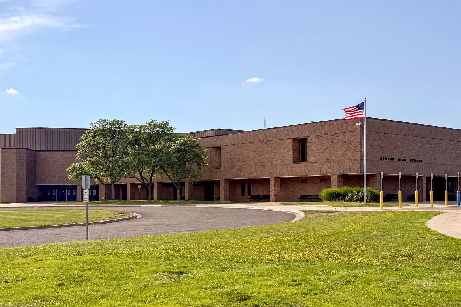 A long, low brown-brick school building with a flagpole and an American flag, behind a curved drive and a wide green lawn under a blue sky, in a file photo.